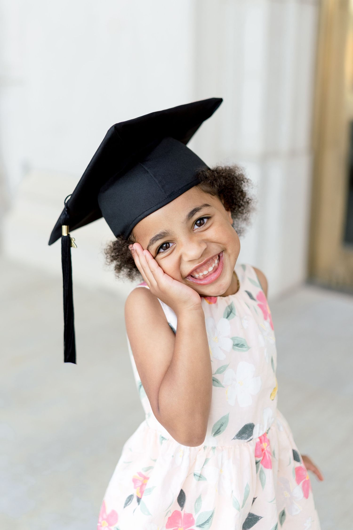 A joyful young girl wearing a black graduation cap and floral dress, smiling with her hand on her cheek.