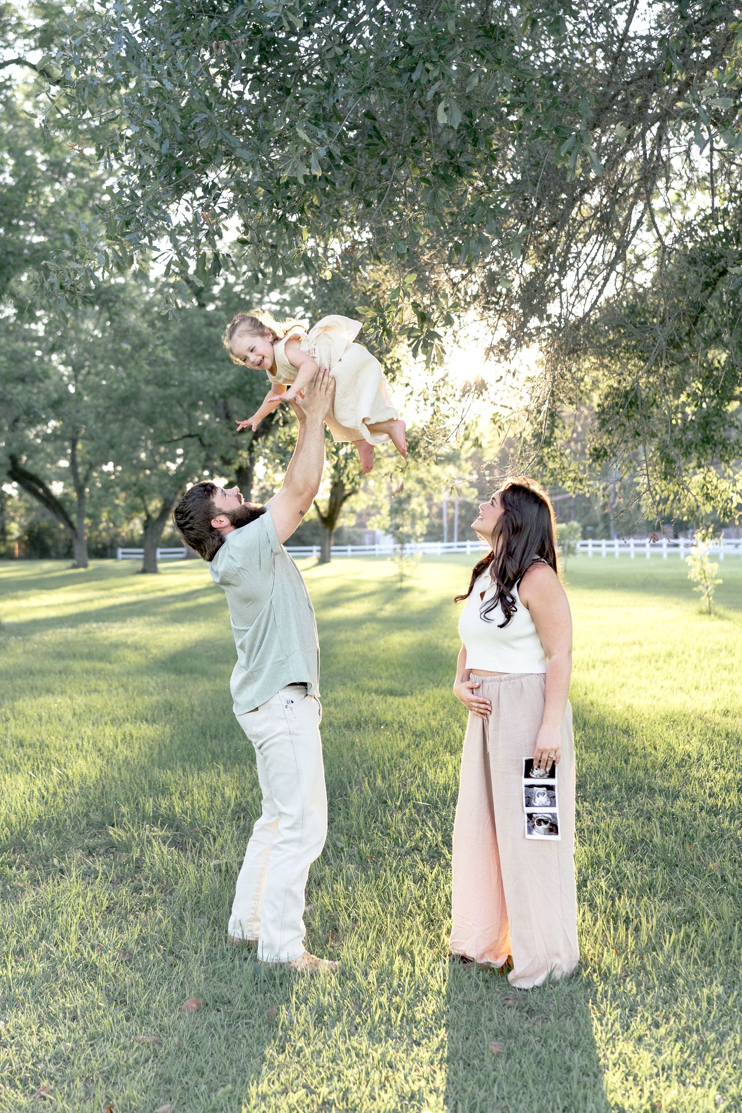 Pregnant woman holds ultrasound photos while man lifts toddler in sunny green park, joyful family maternity announcement photo.