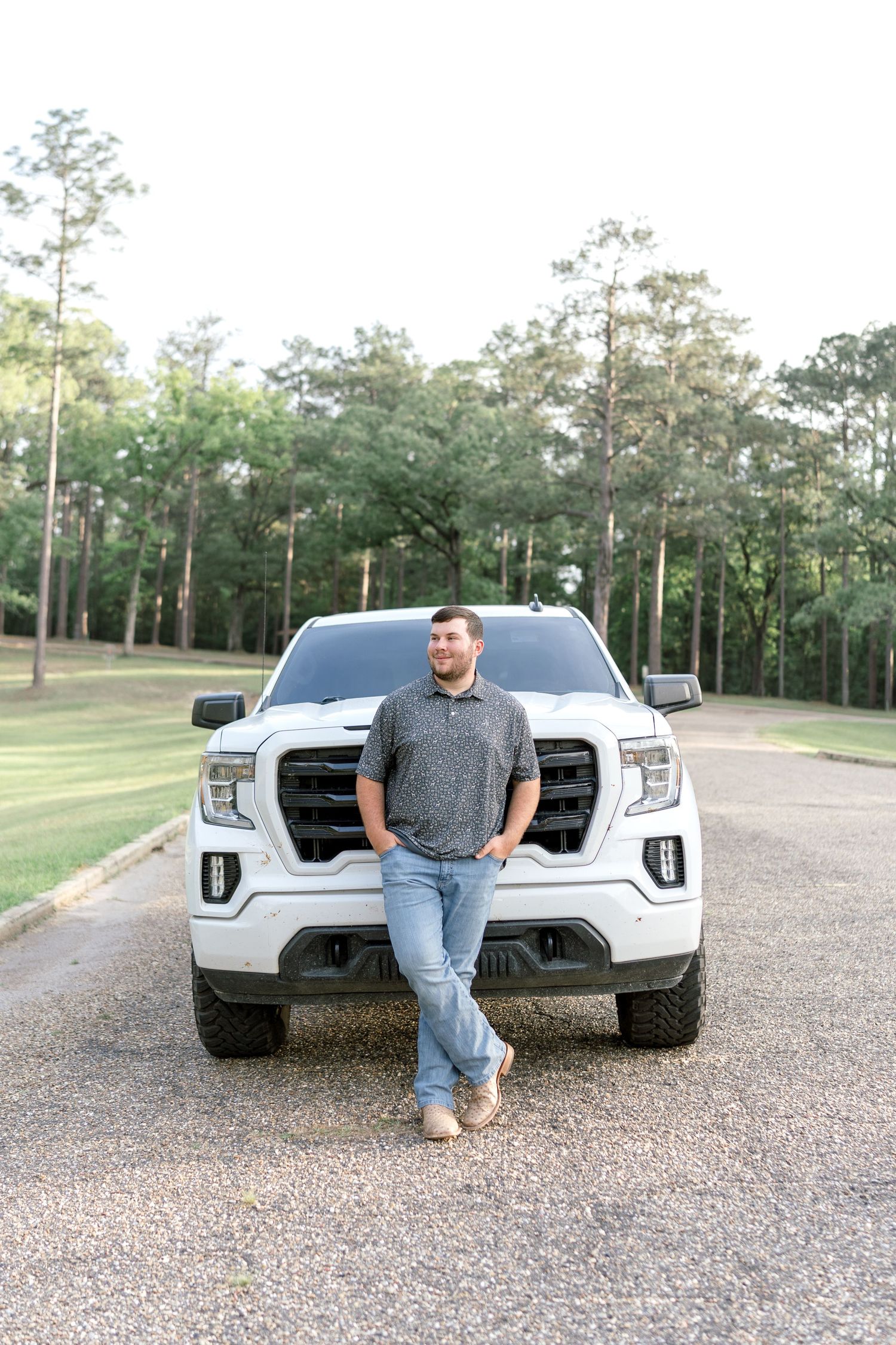 Young man in gray polo and jeans leaning against white GMC truck on gravel driveway surrounded by tall pine trees.
