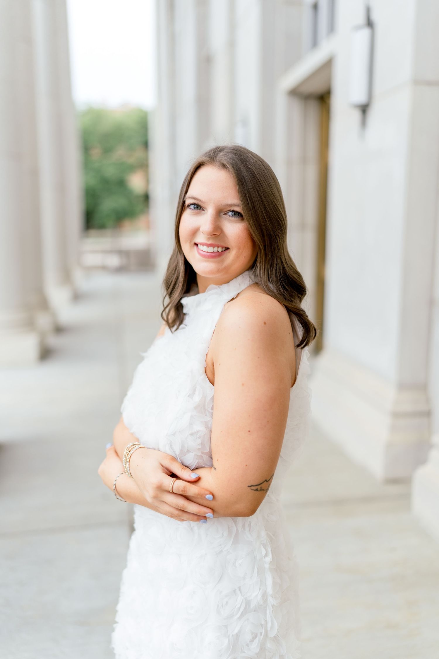 A smiling young woman in a white feathered dress poses with arms crossed near elegant stone columns outdoors.