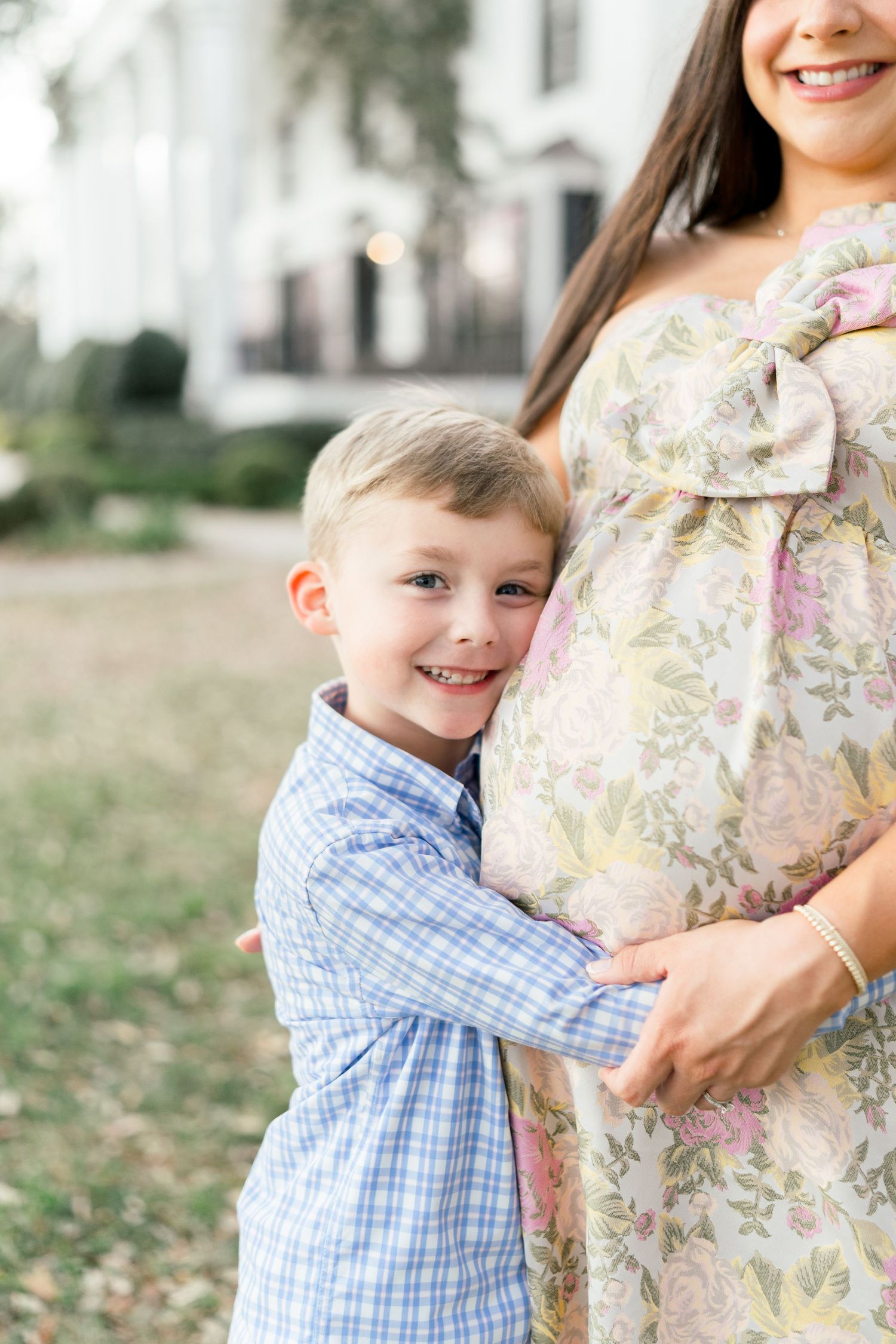 A young boy in a blue plaid shirt hugs a pregnant woman in a floral dress outdoors, smiling at the camera.