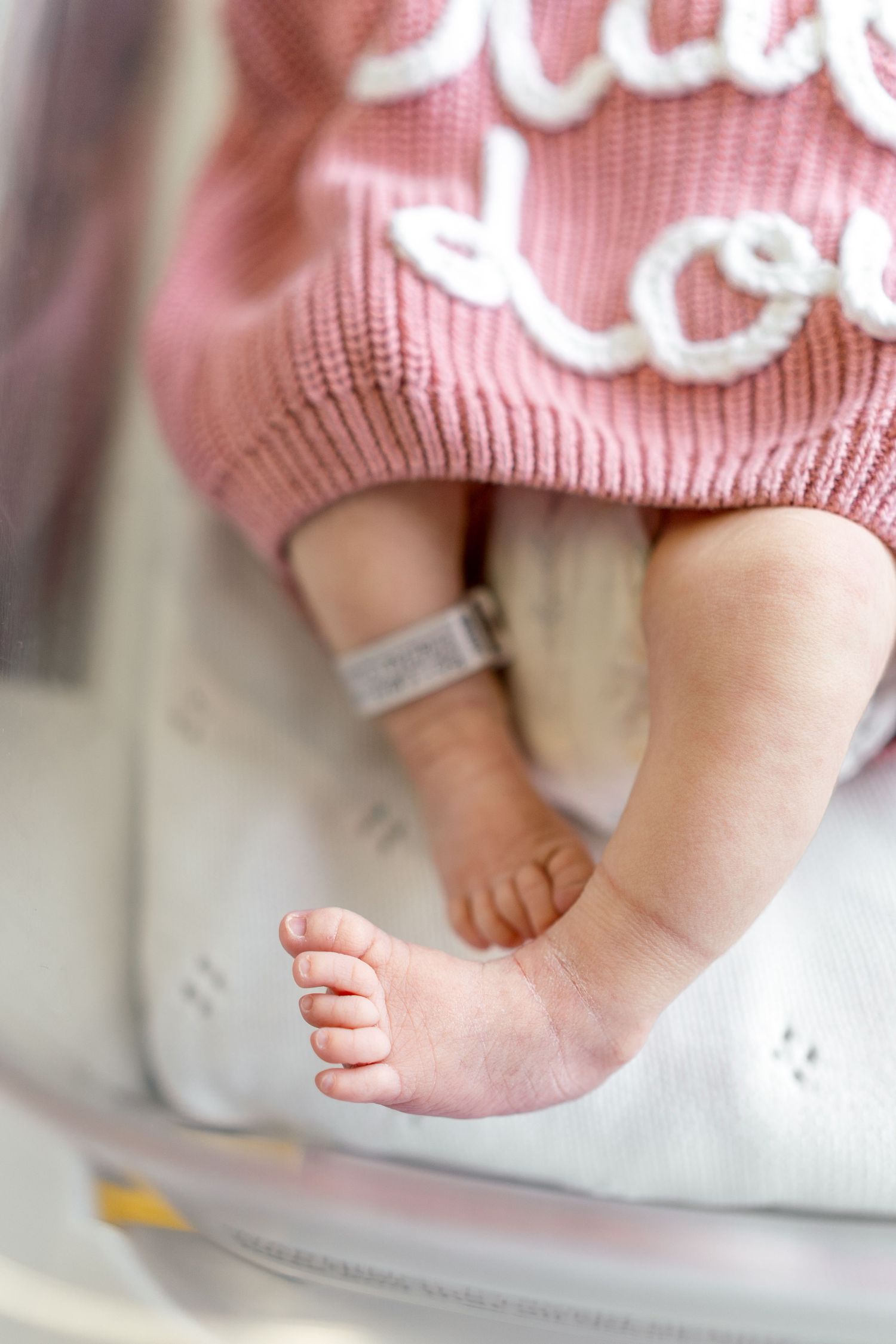 Close-up of newborn baby feet wearing a pink knit outfit with an identification bracelet on a white blanket.