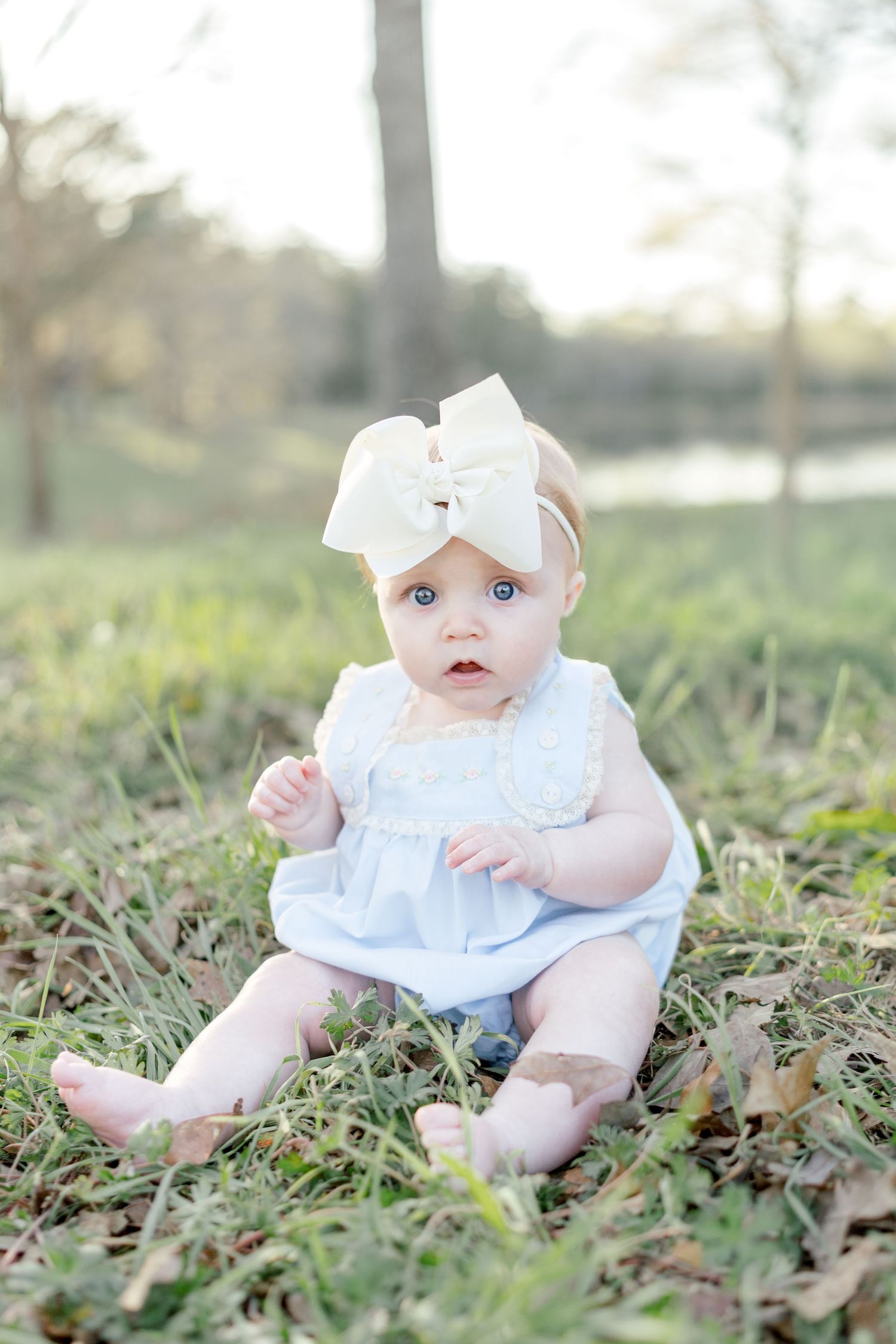 A baby girl in a light blue dress and large cream bow headband sits on grass outdoors in a soft natural setting.