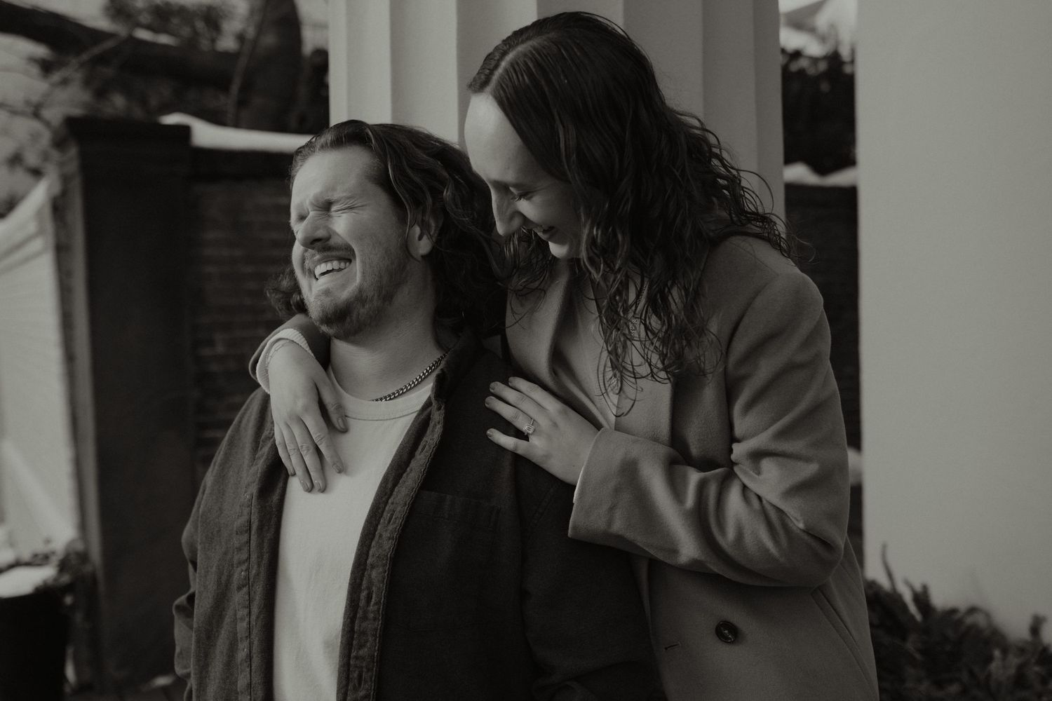 Two men with long hair laugh together outdoors in a black and white photo, sharing a joyful moment.