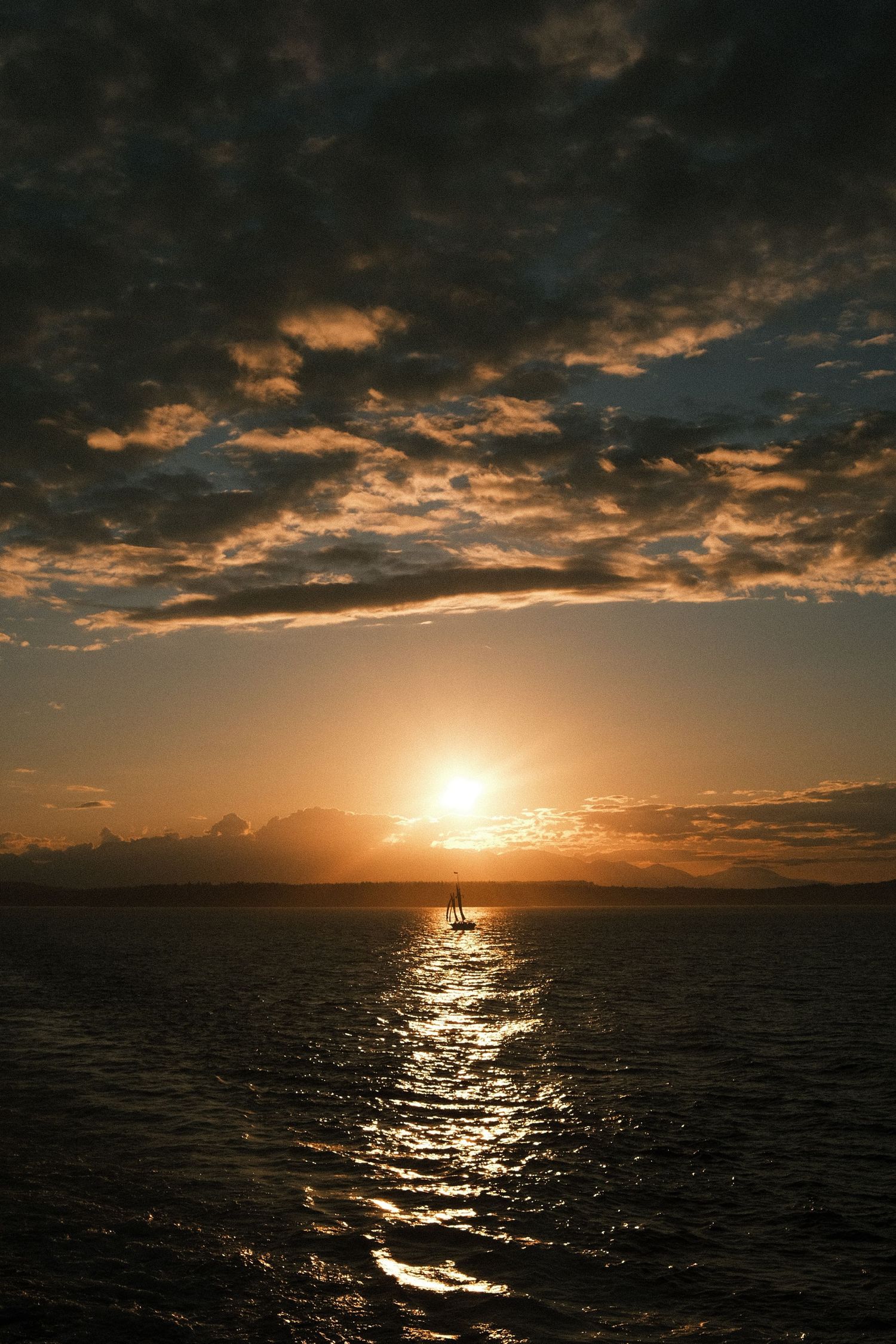 Golden sunset over calm ocean waters with dramatic cloudy sky and silhouetted sailboat reflecting on the sea.