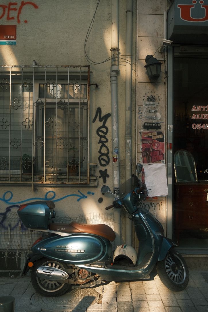Vintage blue scooter parked against a graffiti-covered wall in a narrow urban alleyway with warm sunlight.
