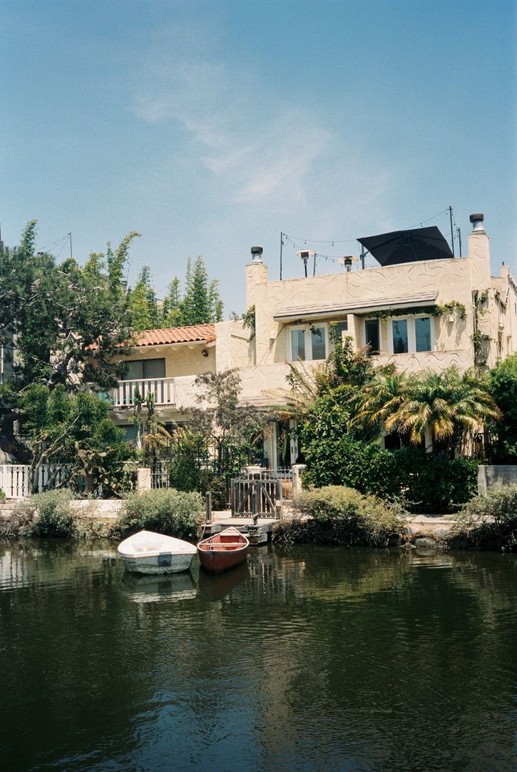 Two small boats moored along a tranquil canal beside a charming beige house surrounded by lush trees.