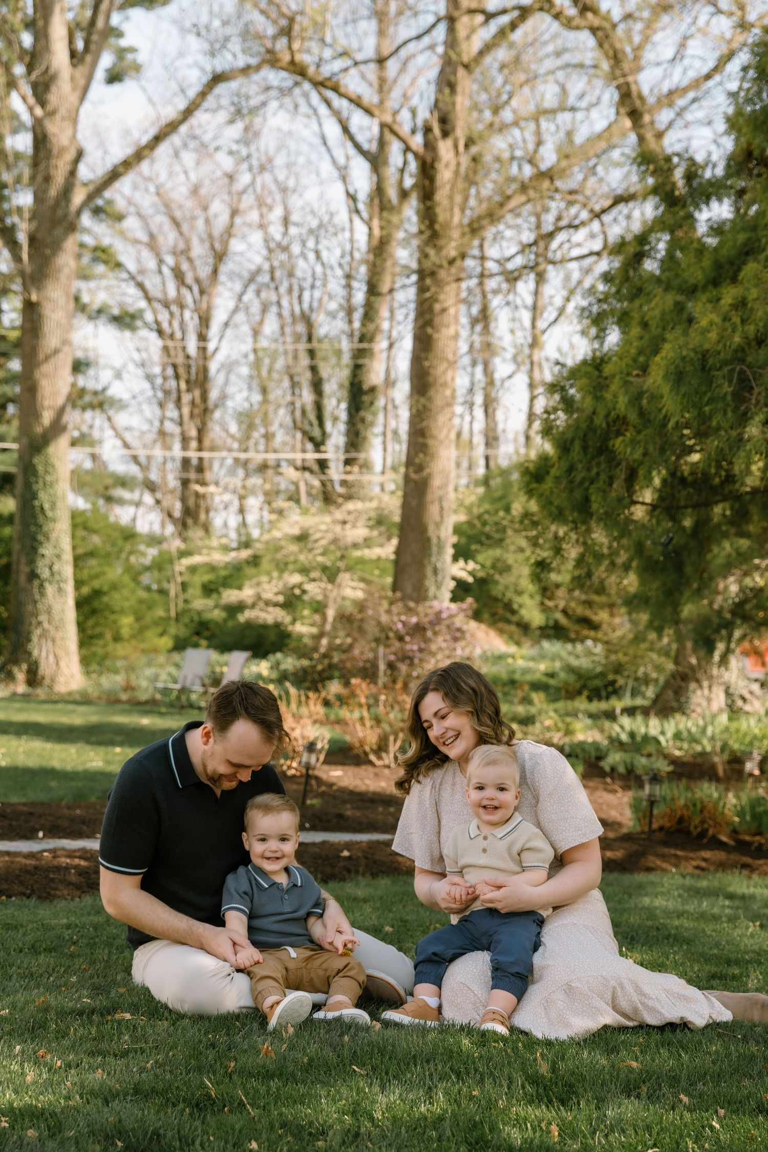 Family of four sitting on grass in a park, parents smiling with two toddlers during a golden hour outdoor photo session.