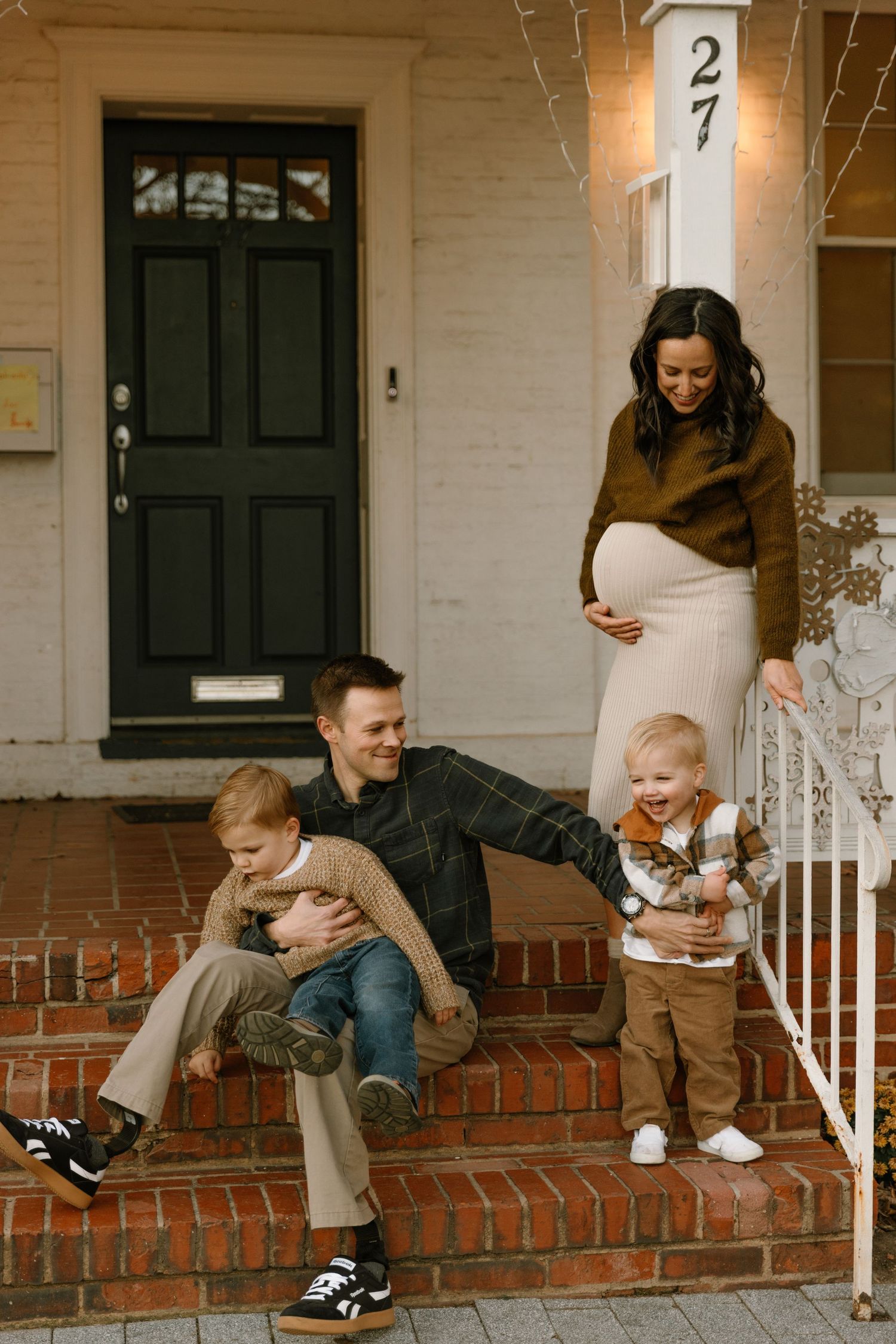 Pregnant woman with husband and two toddler boys sitting on brick front porch steps of white home.
