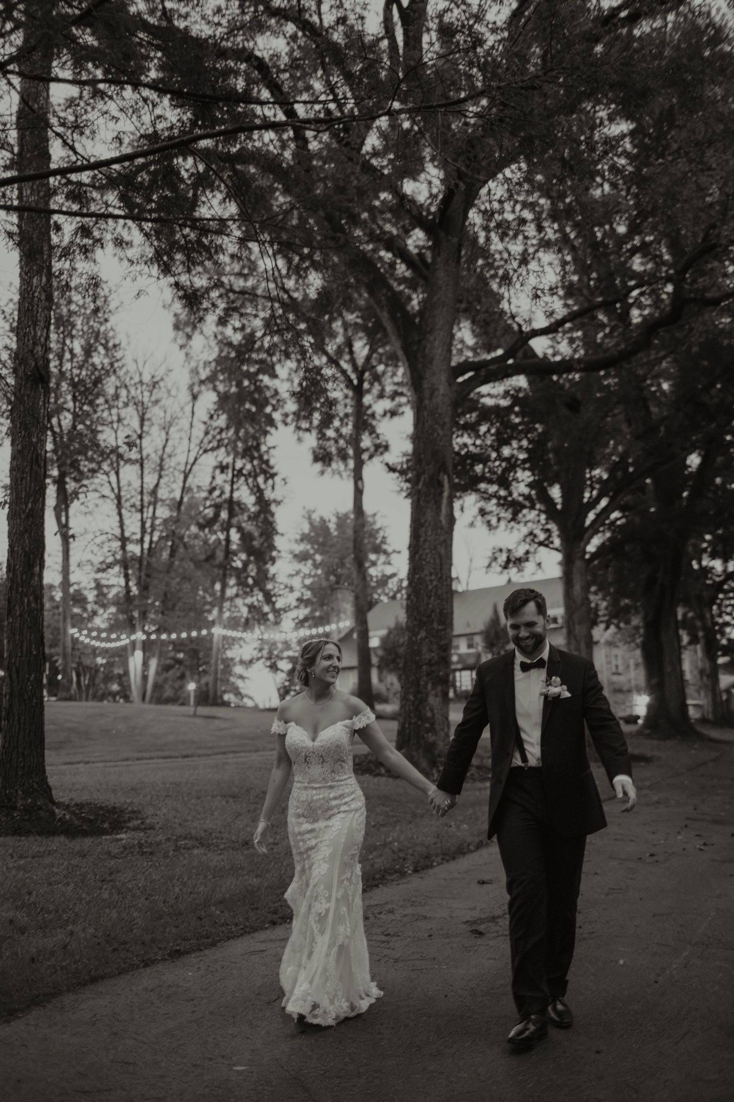 Black and white photo of a bride and groom holding hands, walking along a tree-lined path on their wedding day.