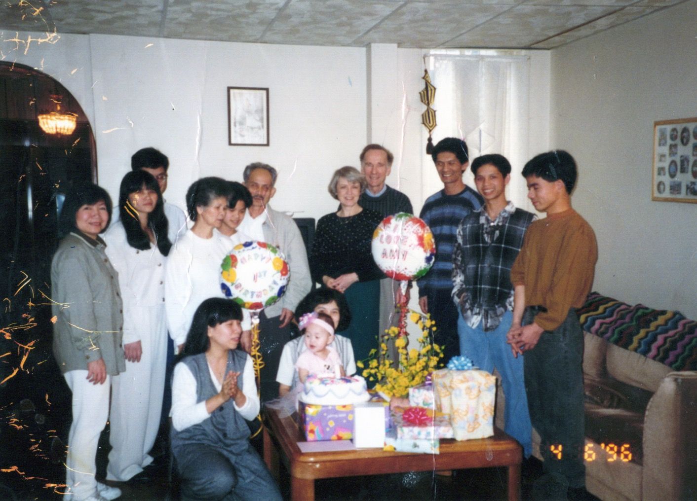 A group of people celebrating a birthday party indoors, holding balloons and gathered around a table with gifts and flowers.
