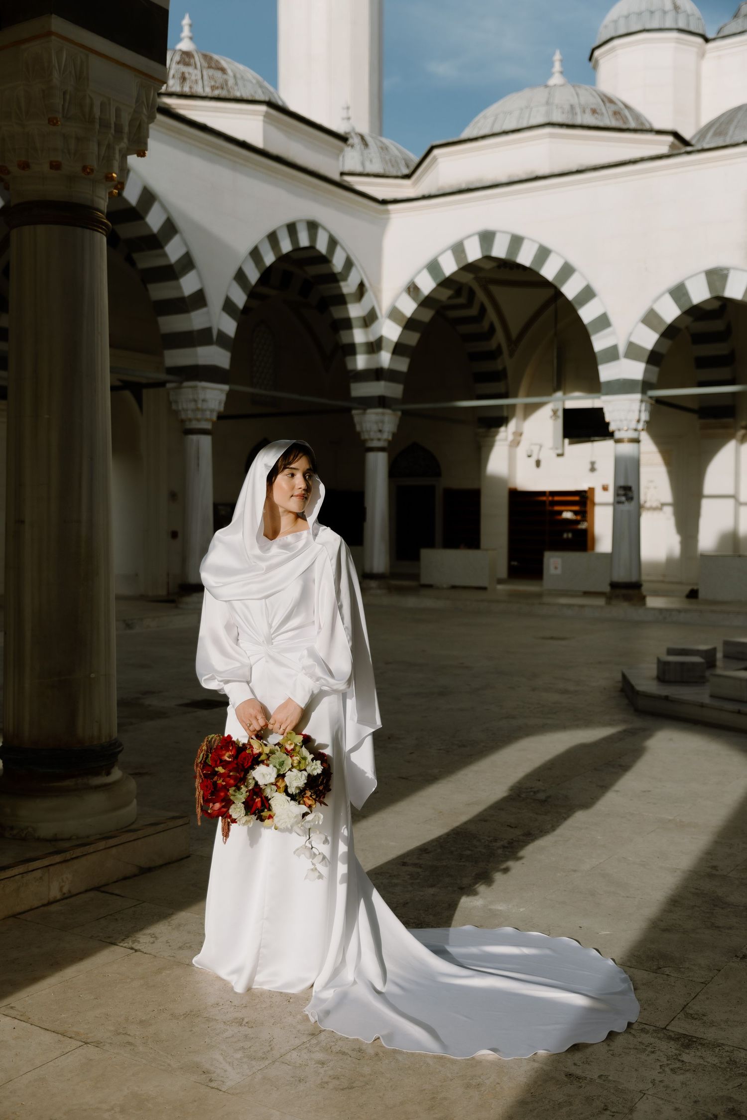 Bride in white Islamic wedding dress holding bouquet in mosque courtyard with arched columns.