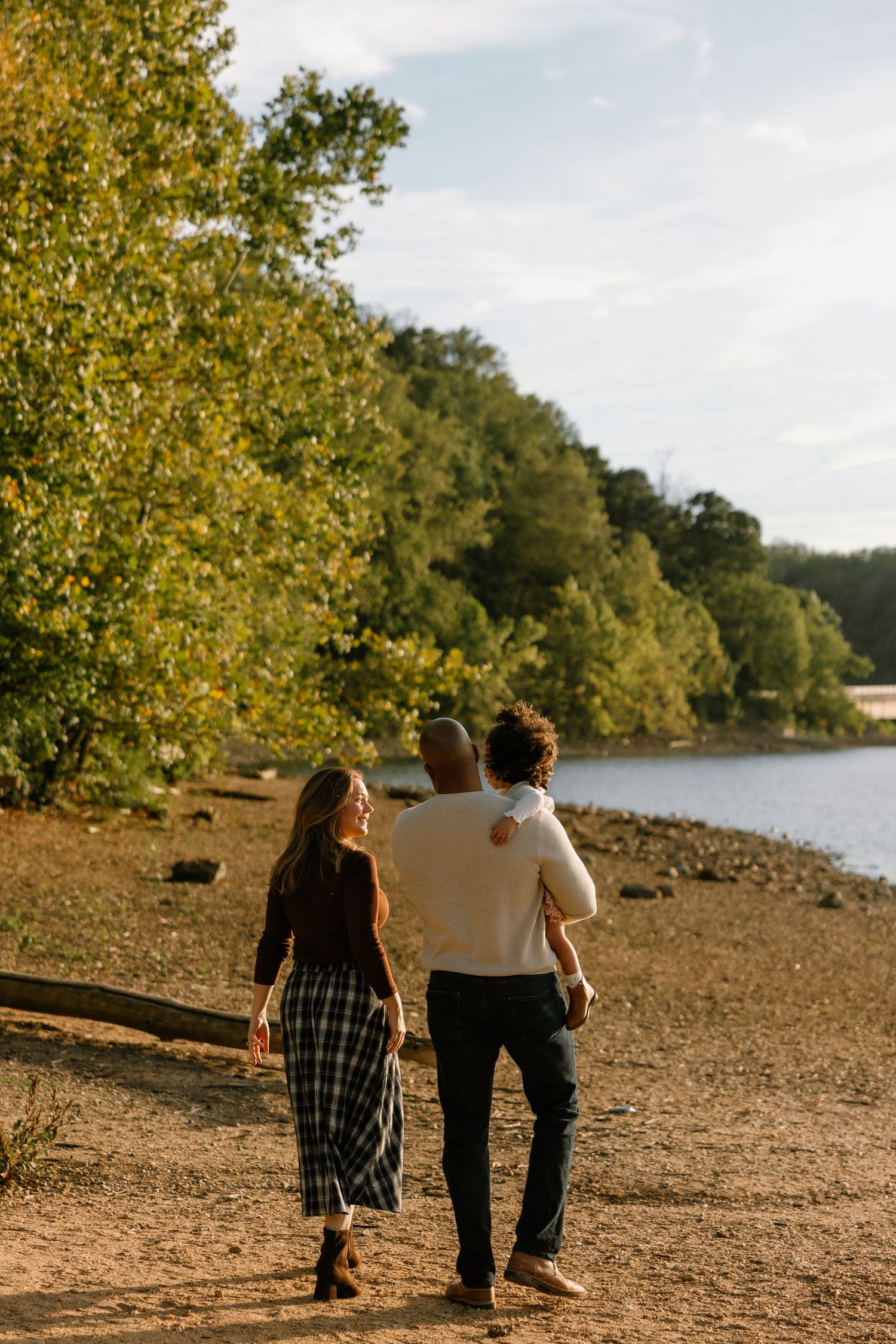 A family walks along a sunny lakeside trail surrounded by lush green trees on a beautiful day.