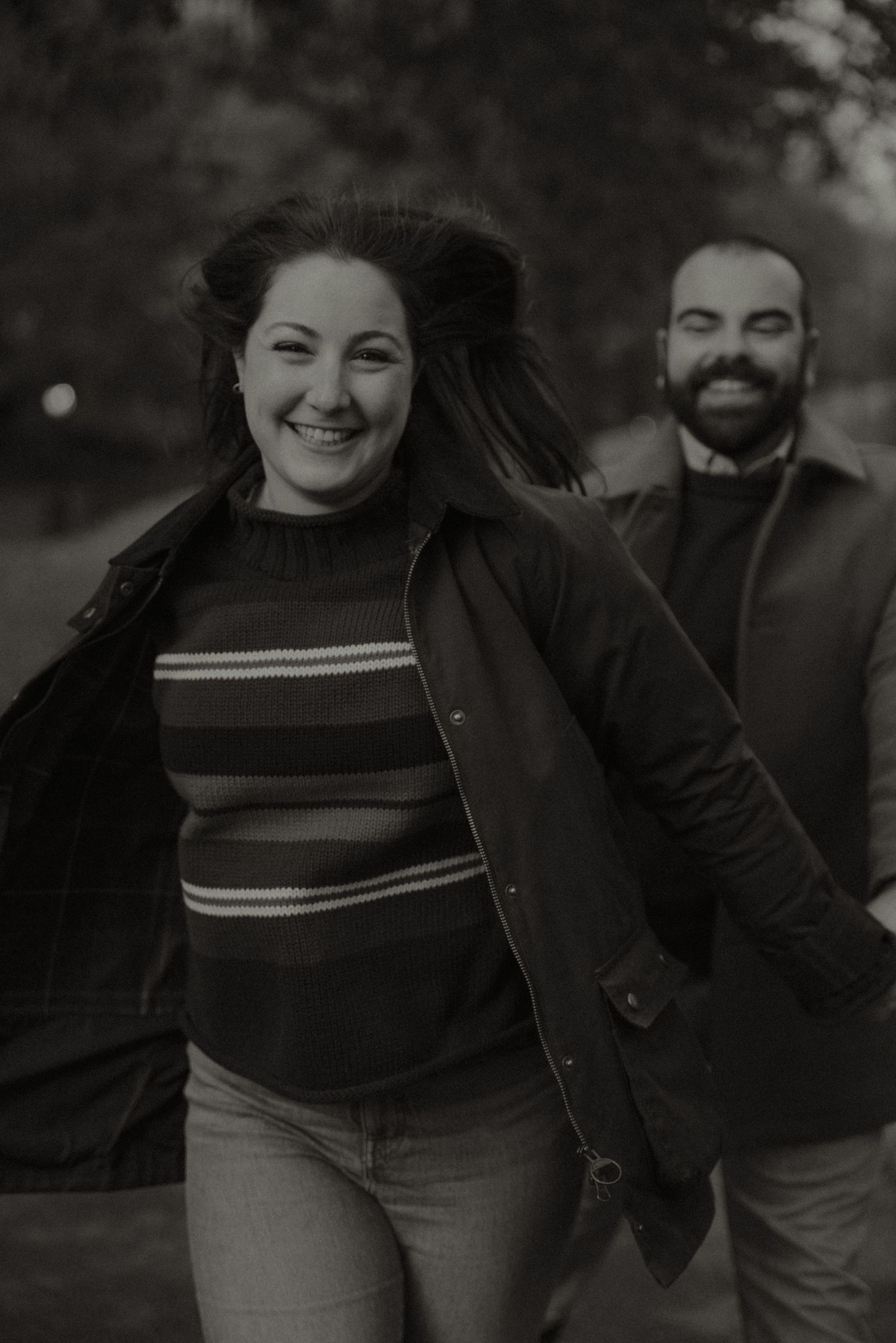 Black and white photo of a smiling woman with windswept hair and a bearded man laughing outdoors.