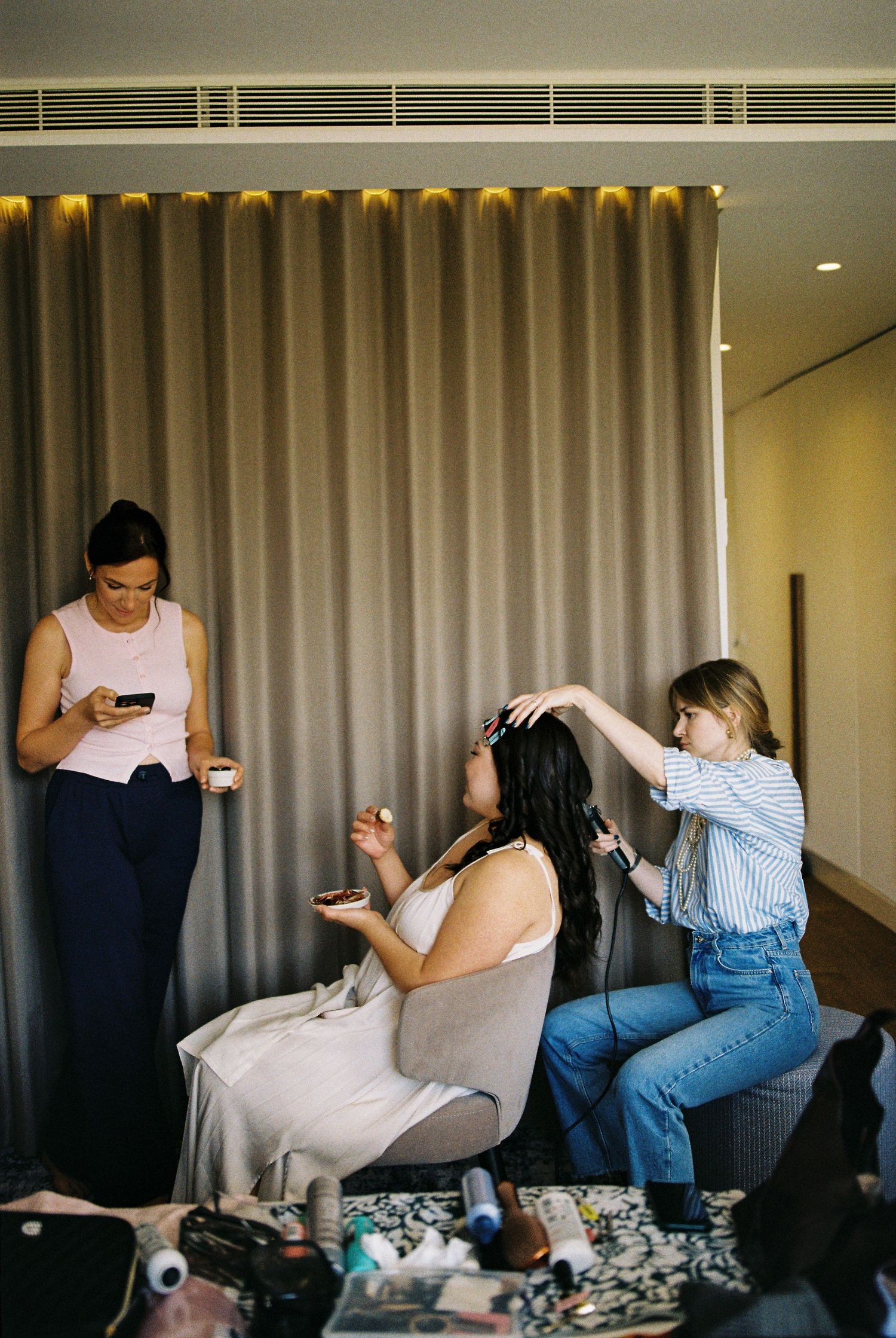 Bride getting hair styled by hairstylist while bridesmaid checks phone in modern hotel room.
