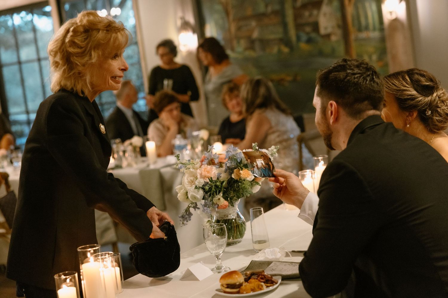 Elegant restaurant dinner scene with guests at candlelit tables, server presenting dish to seated man in black suit.