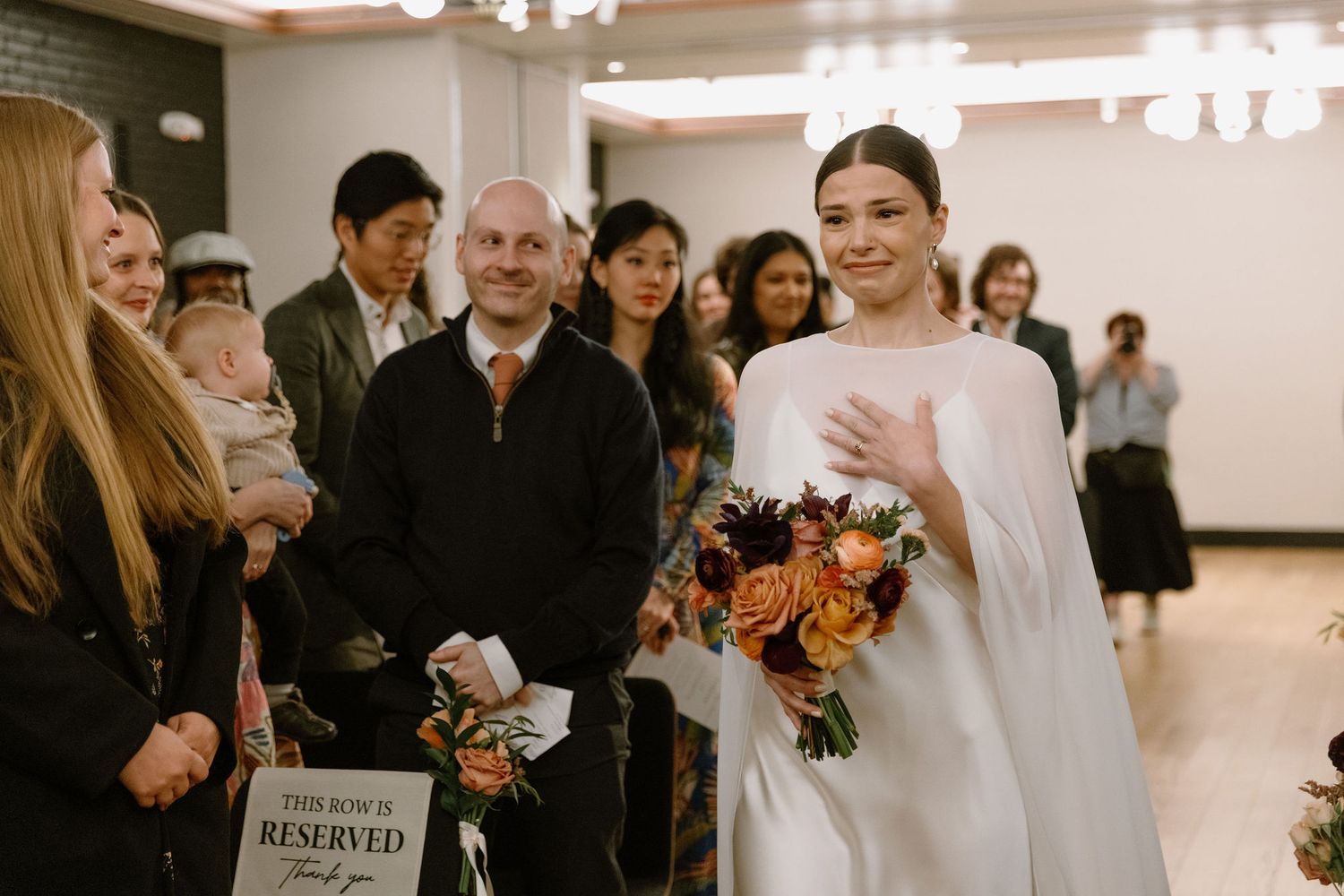 Bride in white cape gown walks down aisle holding colorful bouquet, guests smiling in warmly lit wedding venue.