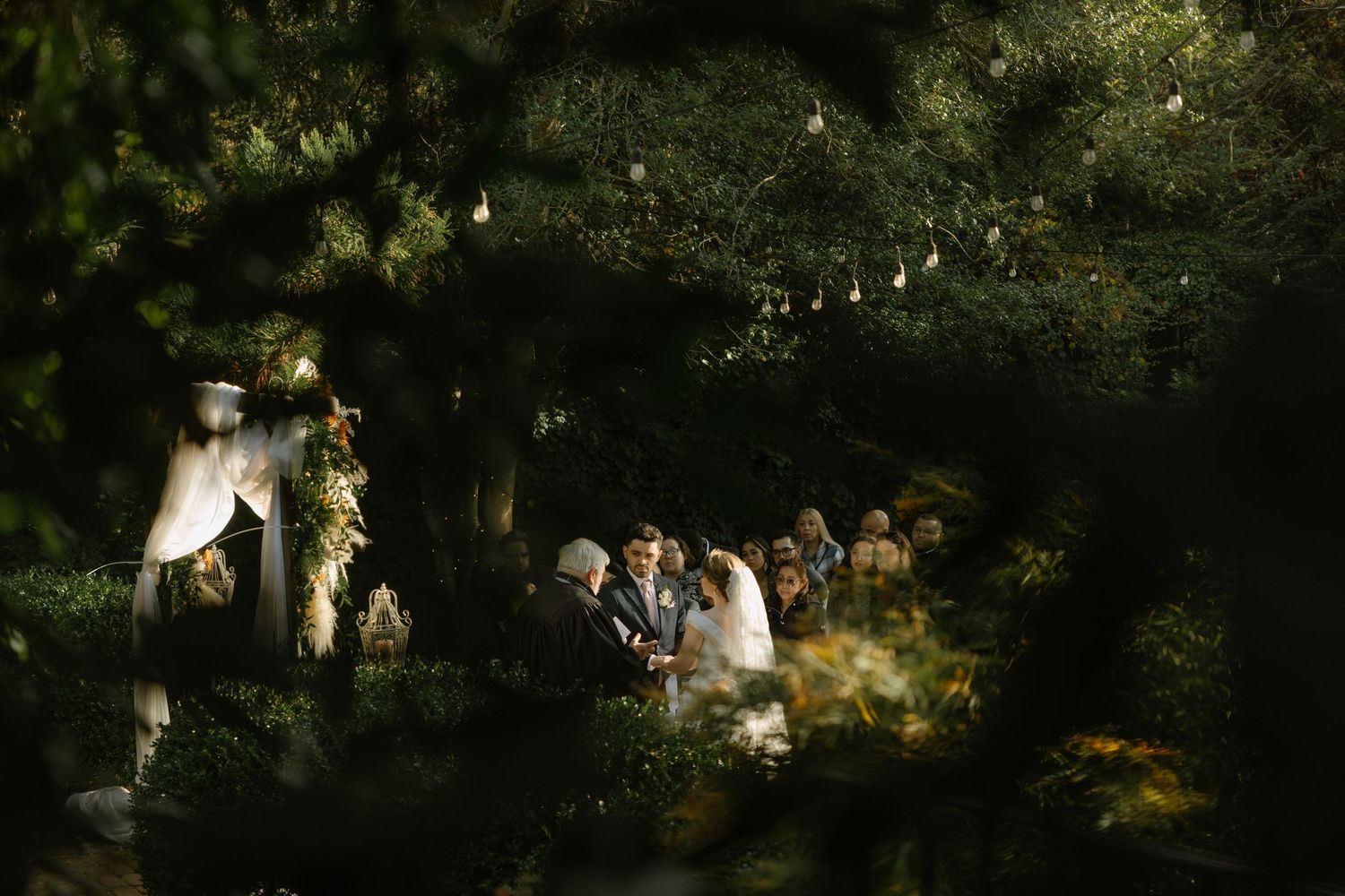 Romantic outdoor wedding ceremony framed by lush green trees with string lights glowing softly in a garden setting.