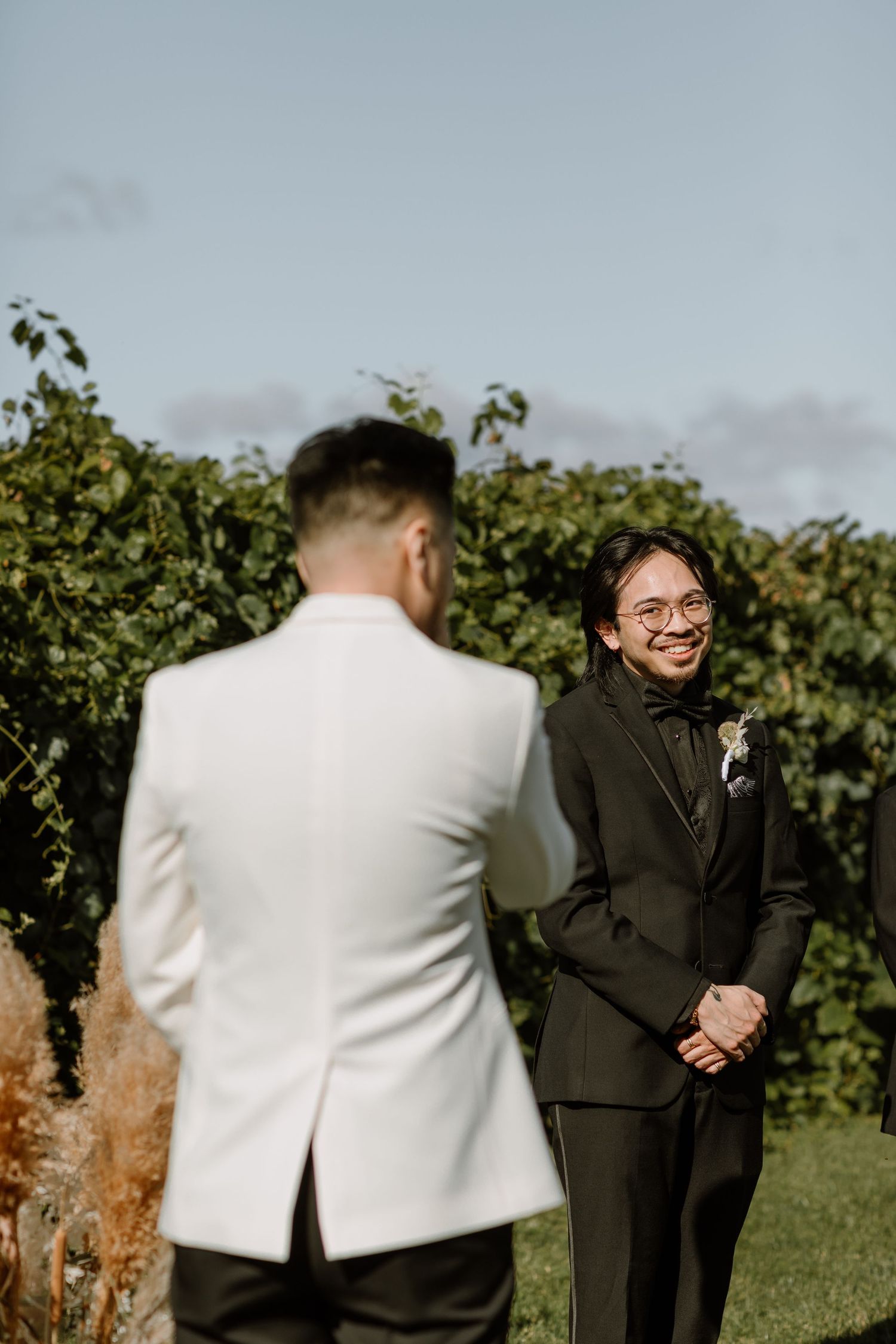 Two grooms share a joyful moment outdoors at a vineyard wedding, one in white and one in a black tuxedo.