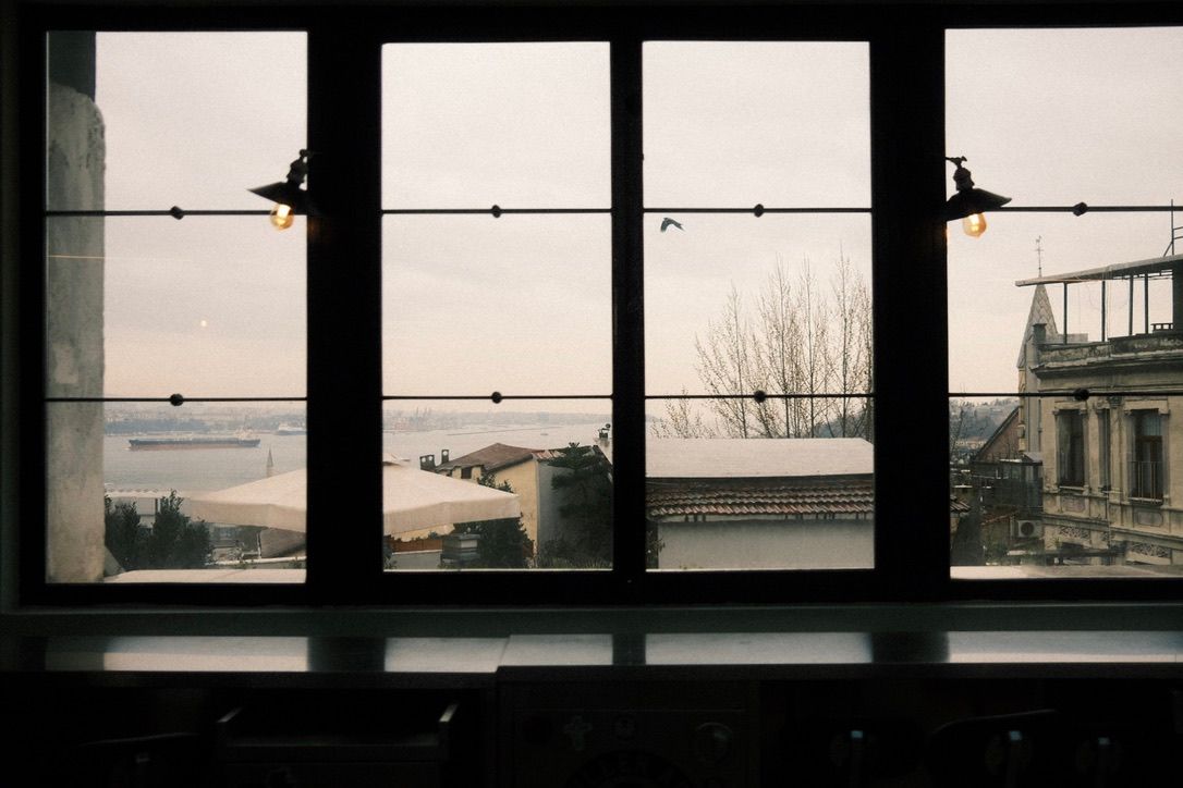 Interior view through large industrial windows overlooking a waterfront cityscape on an overcast day.