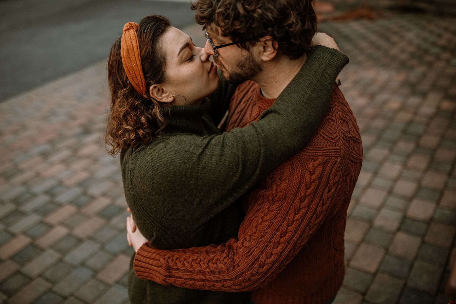 A couple shares a romantic kiss outdoors, embracing warmly in cozy autumn sweaters on a brick pathway.