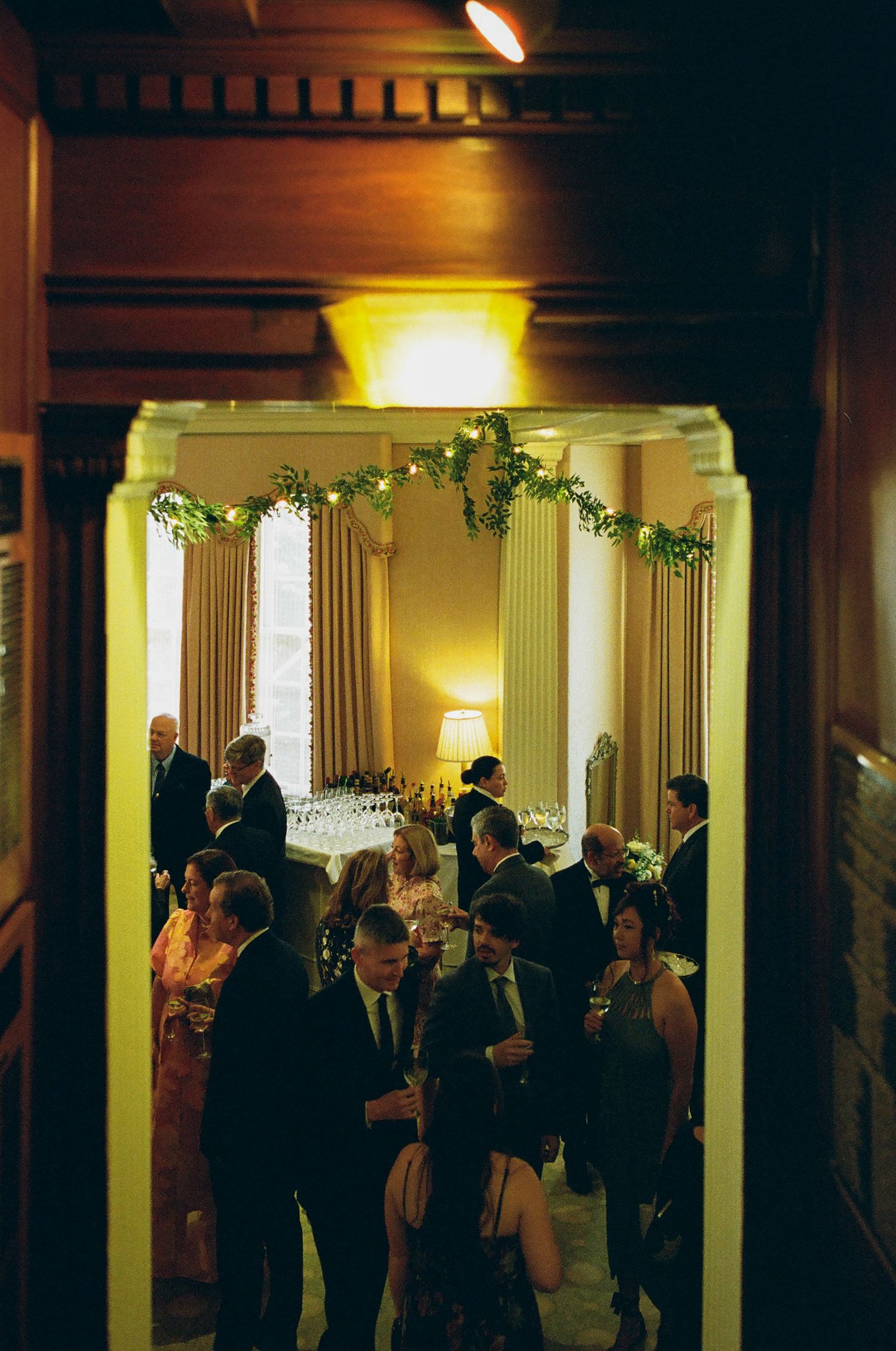 Elegant indoor wedding reception with guests mingling under greenery garland archway in warmly lit venue.