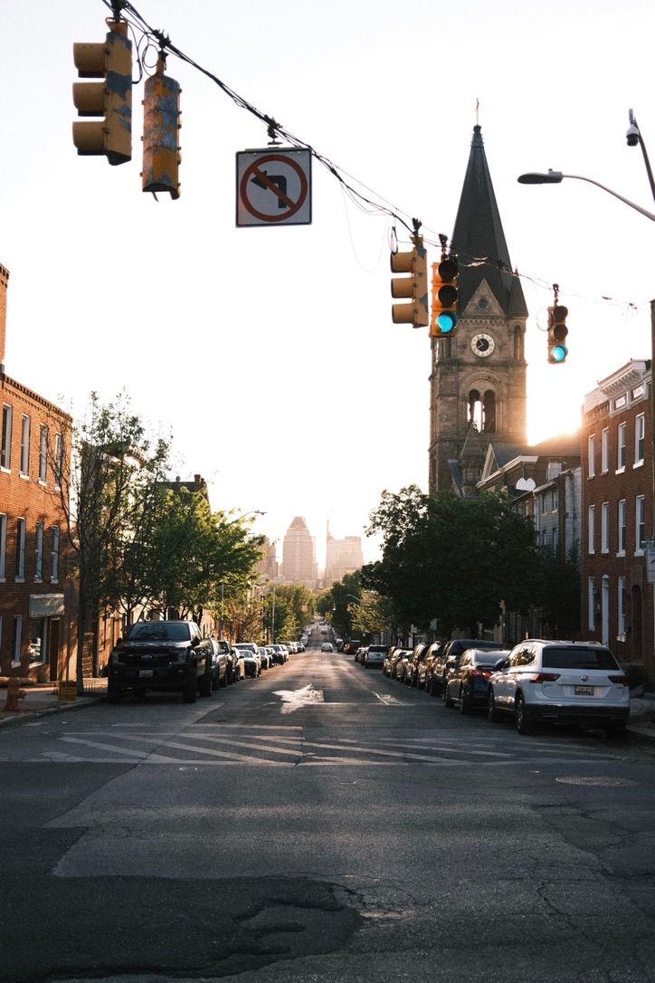 Sunlit urban street with church clock tower, traffic lights, and parked cars lining brick buildings at golden hour.