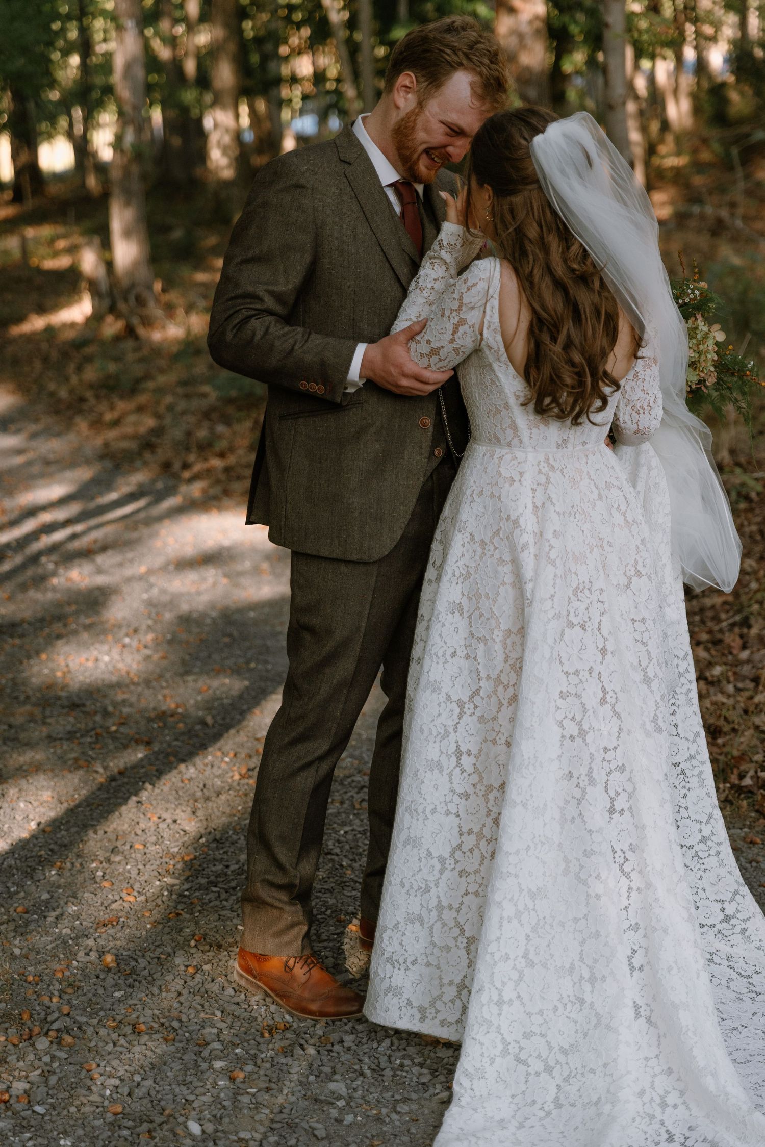 Bride in lace gown and veil embraces groom in brown suit among forest trees on their wedding day.