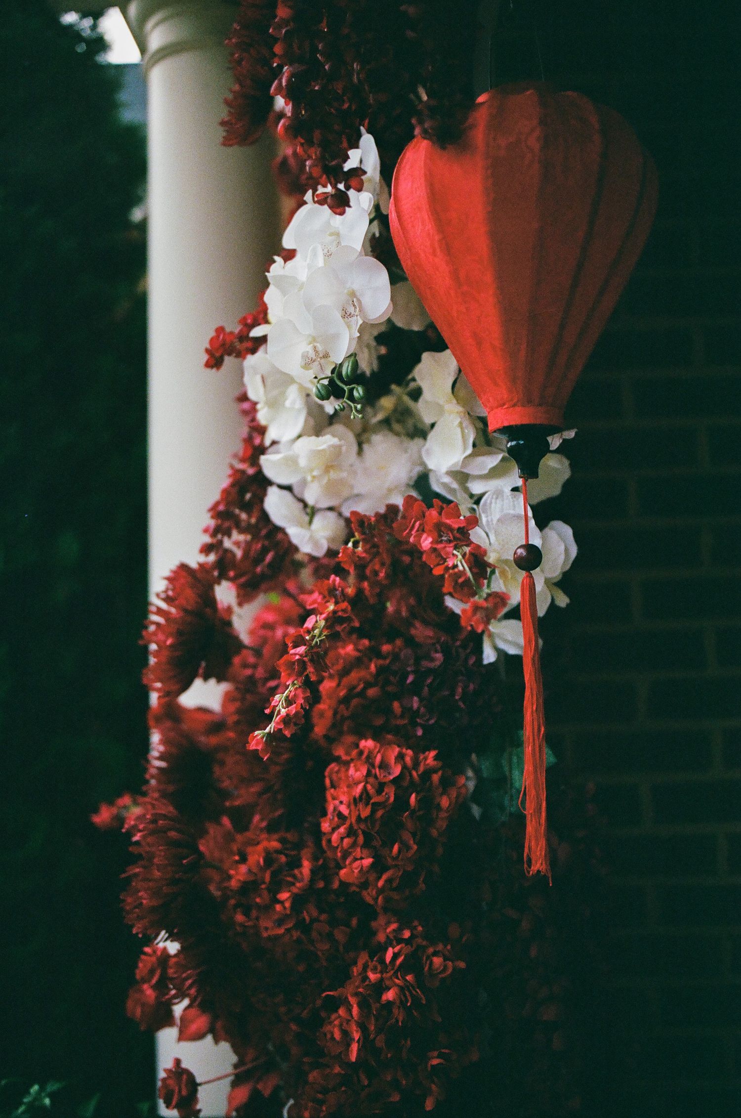 Red Chinese lantern with white and red floral decorations hanging near a white column and brick wall.