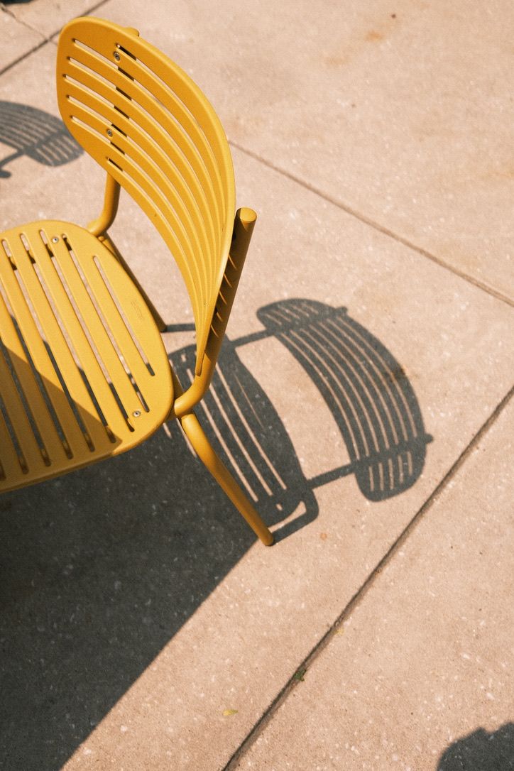 A bright yellow metal slatted chair casting a striped shadow on a beige concrete pavement in sunlight.