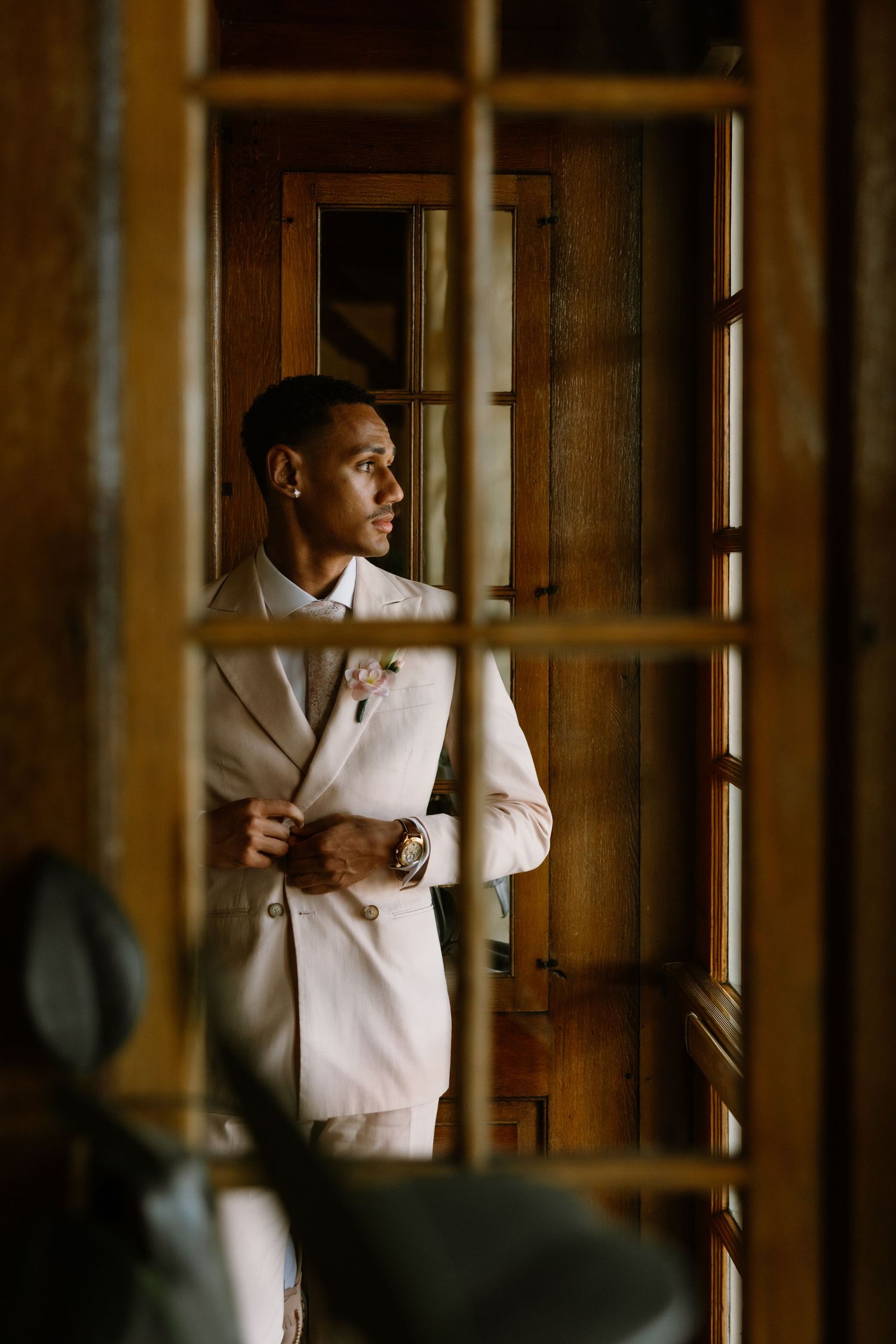 Elegant groom in a white suit adjusting his jacket while gazing out a wooden window.