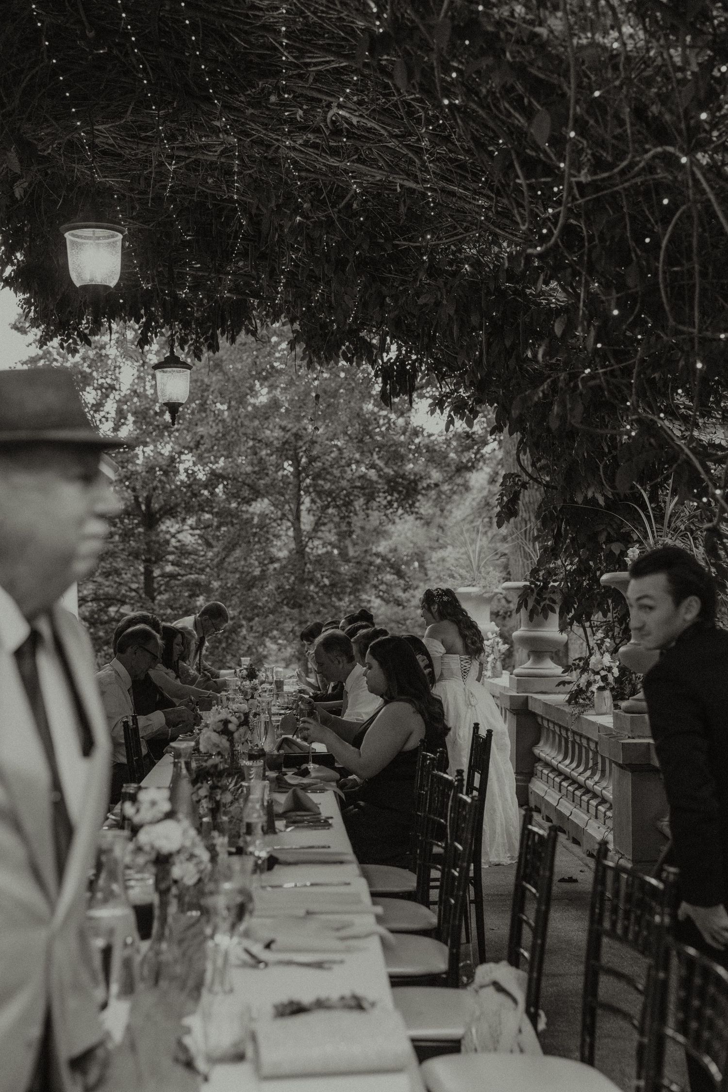 Black and white photo of elegant outdoor wedding reception with long dining table, fairy lights, and guests seated under lush trees.