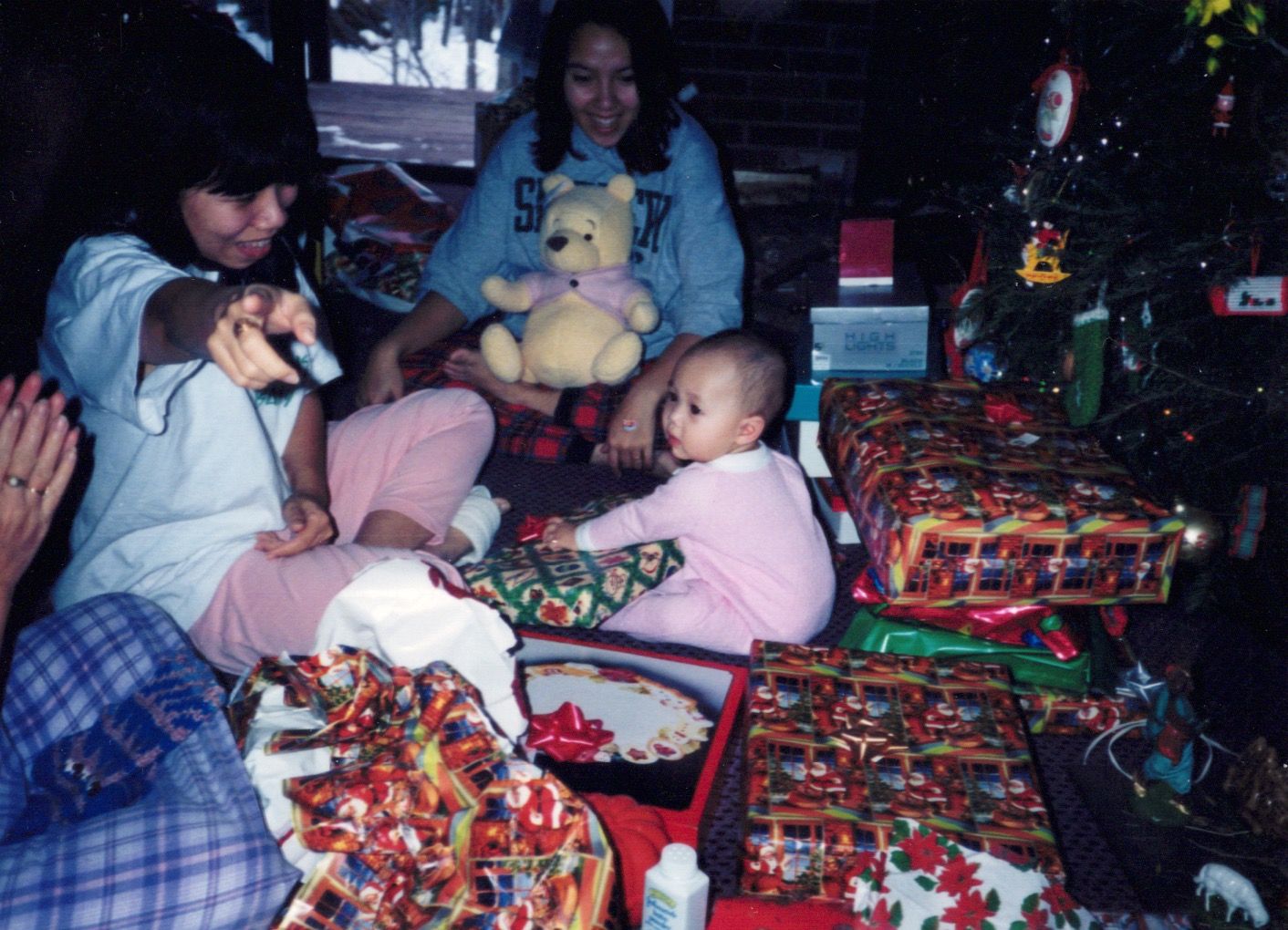 Family celebrating Christmas with wrapped gifts, a baby, and a Winnie the Pooh stuffed animal near a decorated tree.