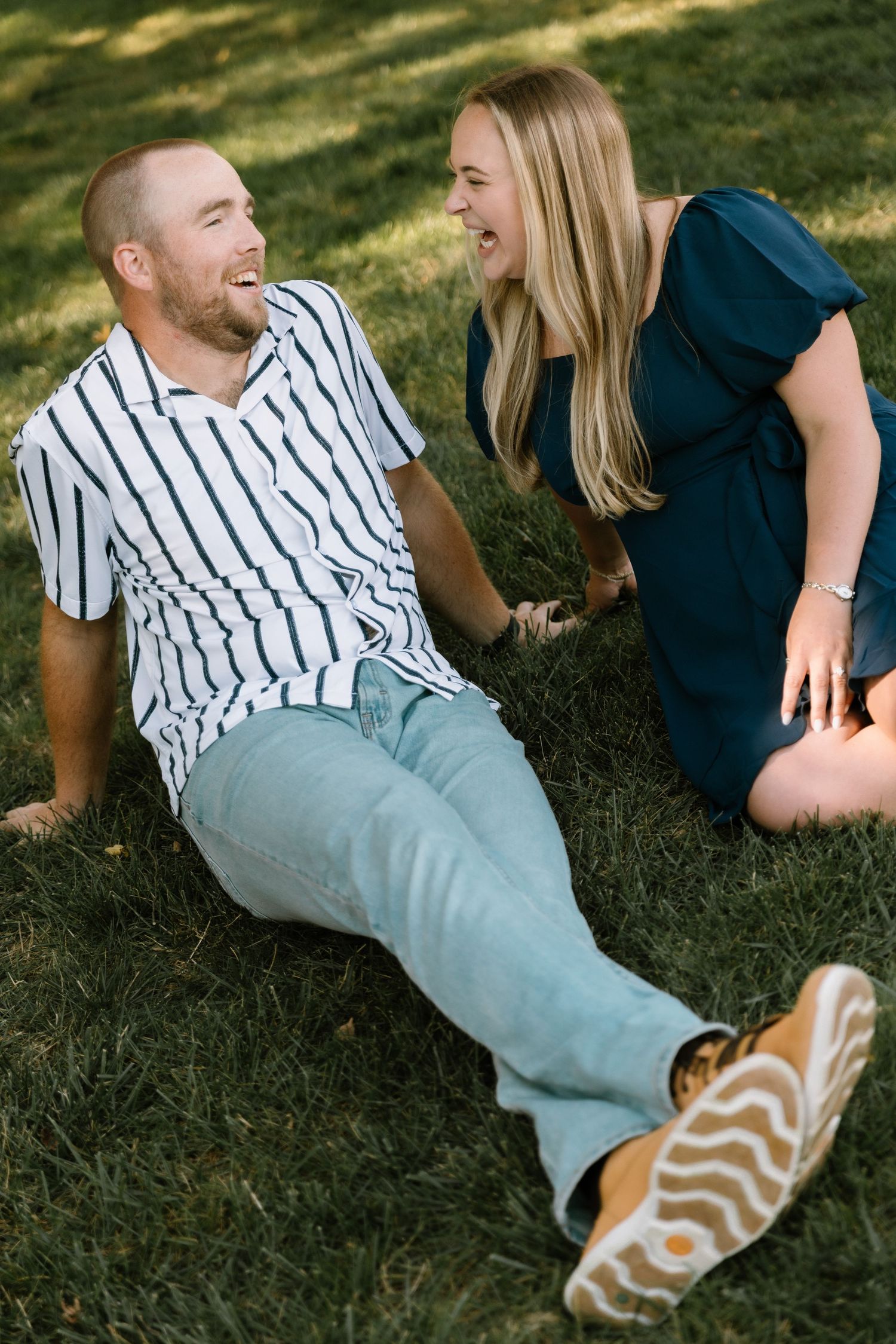 A laughing couple sits on green grass, man in striped shirt and jeans, woman in teal dress, sharing a joyful moment.