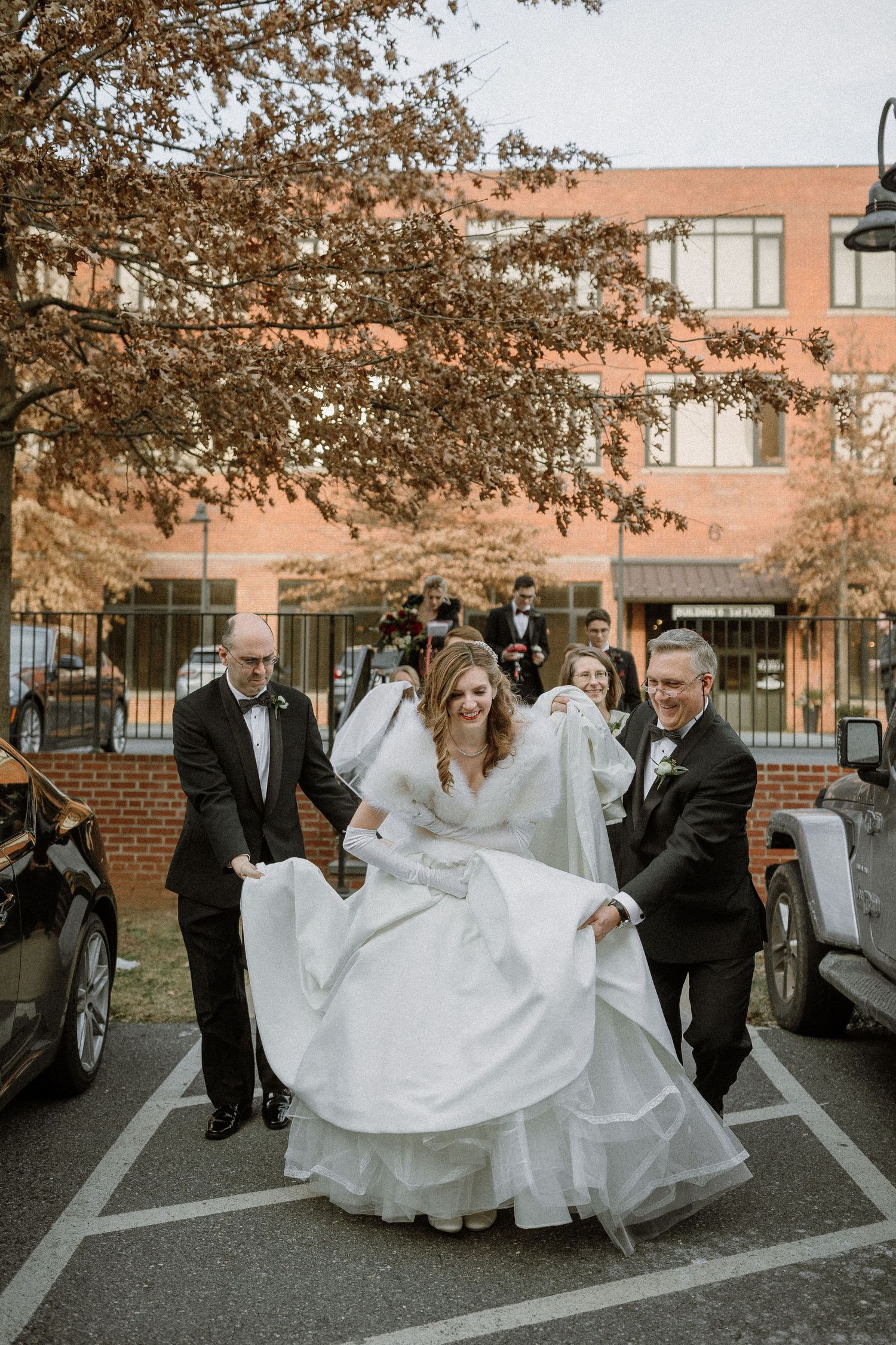 Bride in white gown assisted by two men in tuxedos walking through parking lot outside brick building on wedding day.