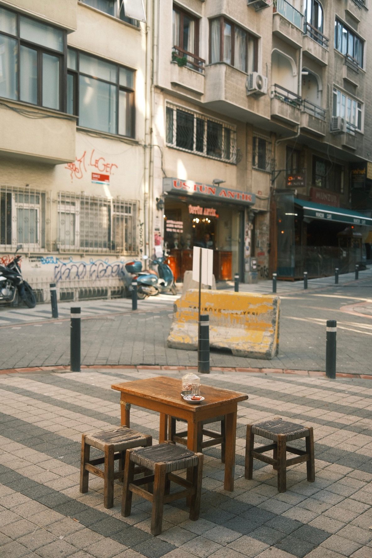 Wooden table with stools on a cobblestone street in Istanbul, with urban buildings and a road barrier in the background.