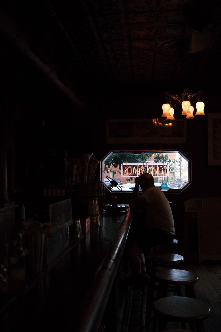 Dimly lit bar interior with a lone patron sitting at the counter near a bright window, warm chandelier glow overhead.