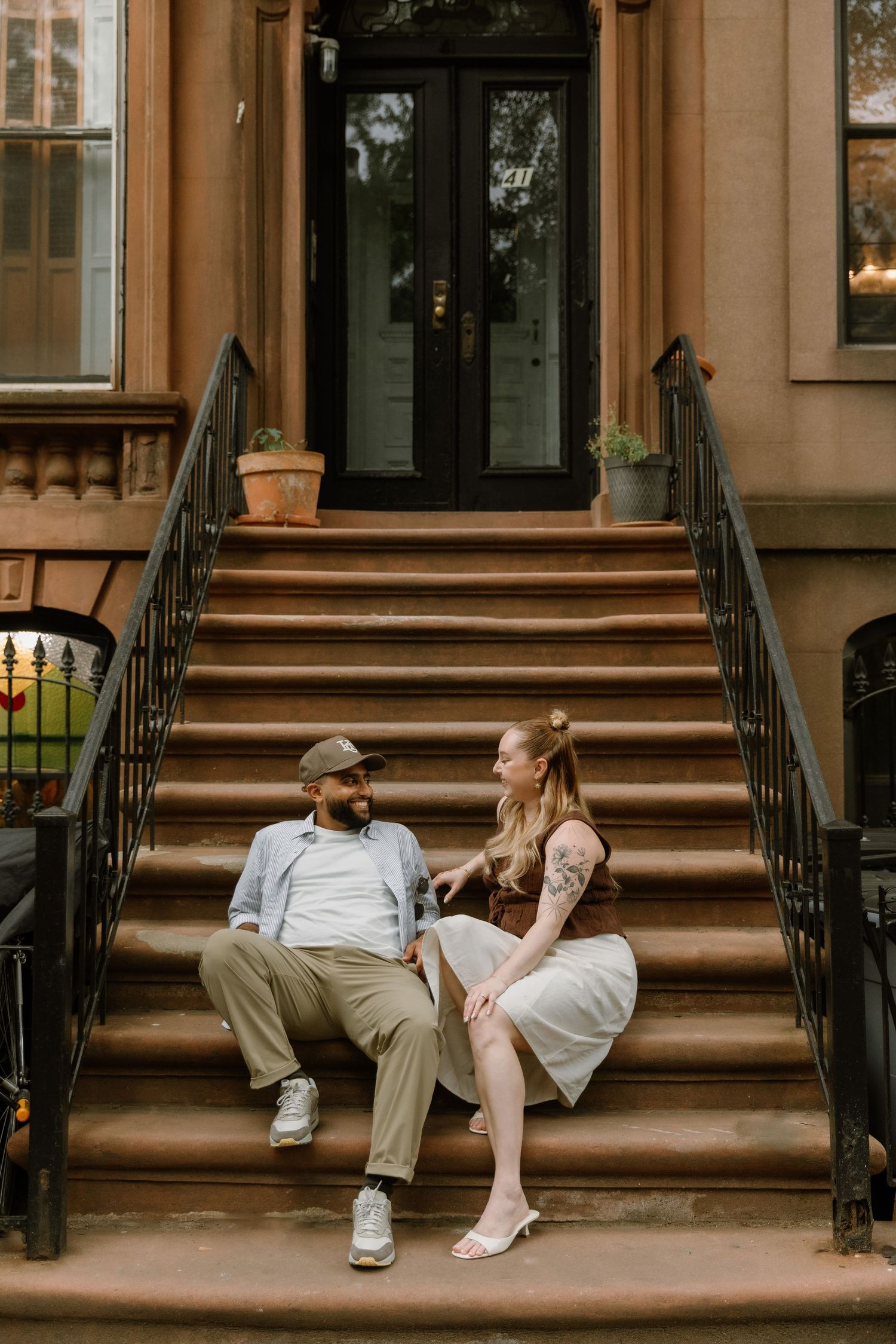 Couple sitting on brownstone stoop steps in NYC, sharing a romantic conversation, woman in skirt and man in casual attire.