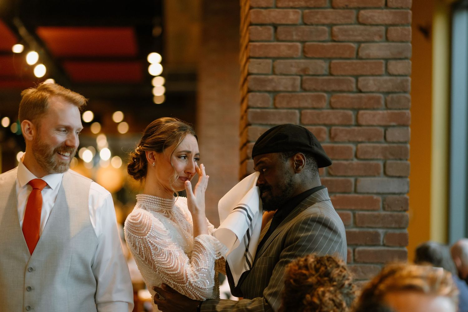 Bride wiping tears at wedding reception near brick pillar, with groom in white suit and guest nearby.