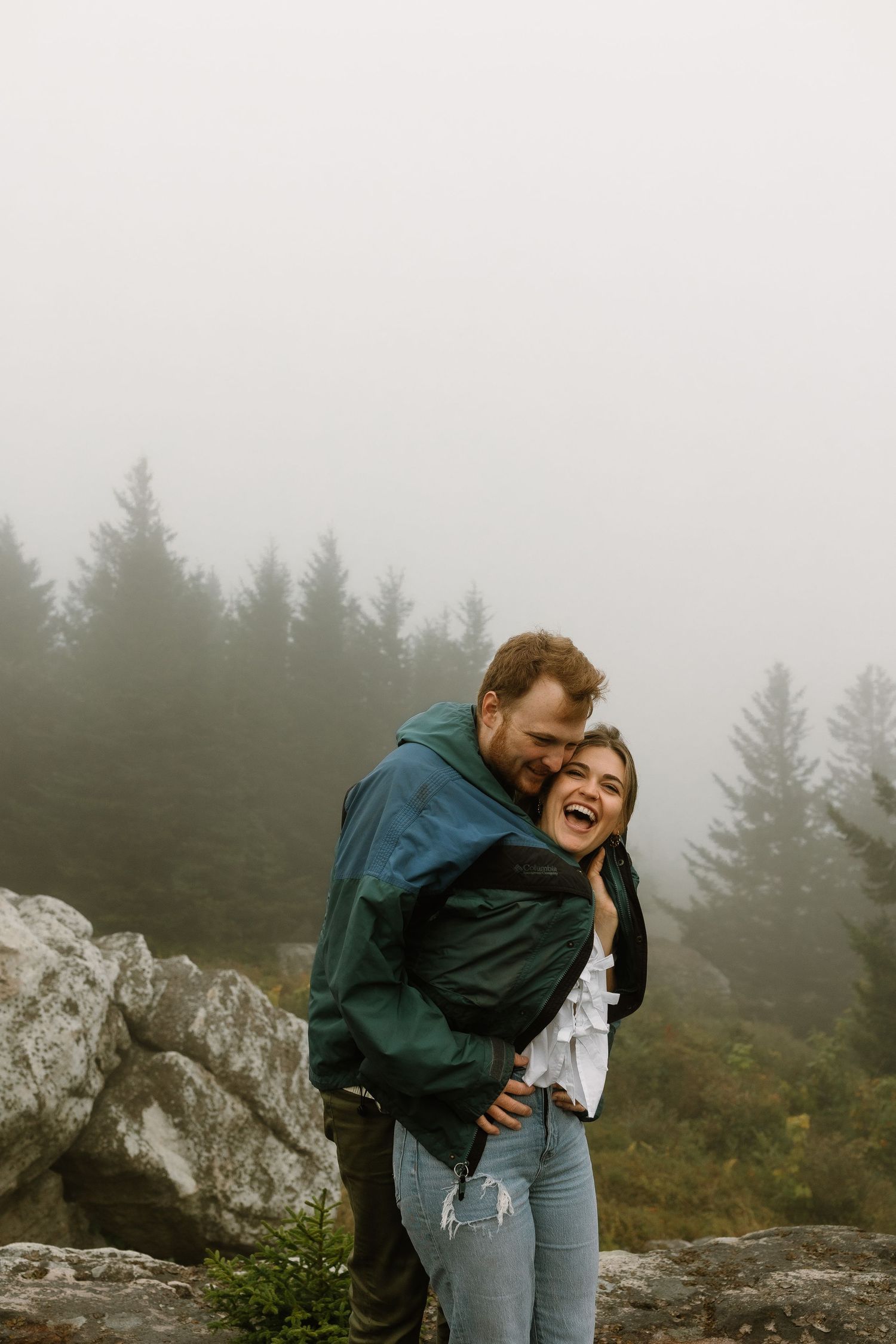 A joyful couple embraces on a rocky overlook surrounded by misty pine trees, sharing a playful and loving moment outdoors.