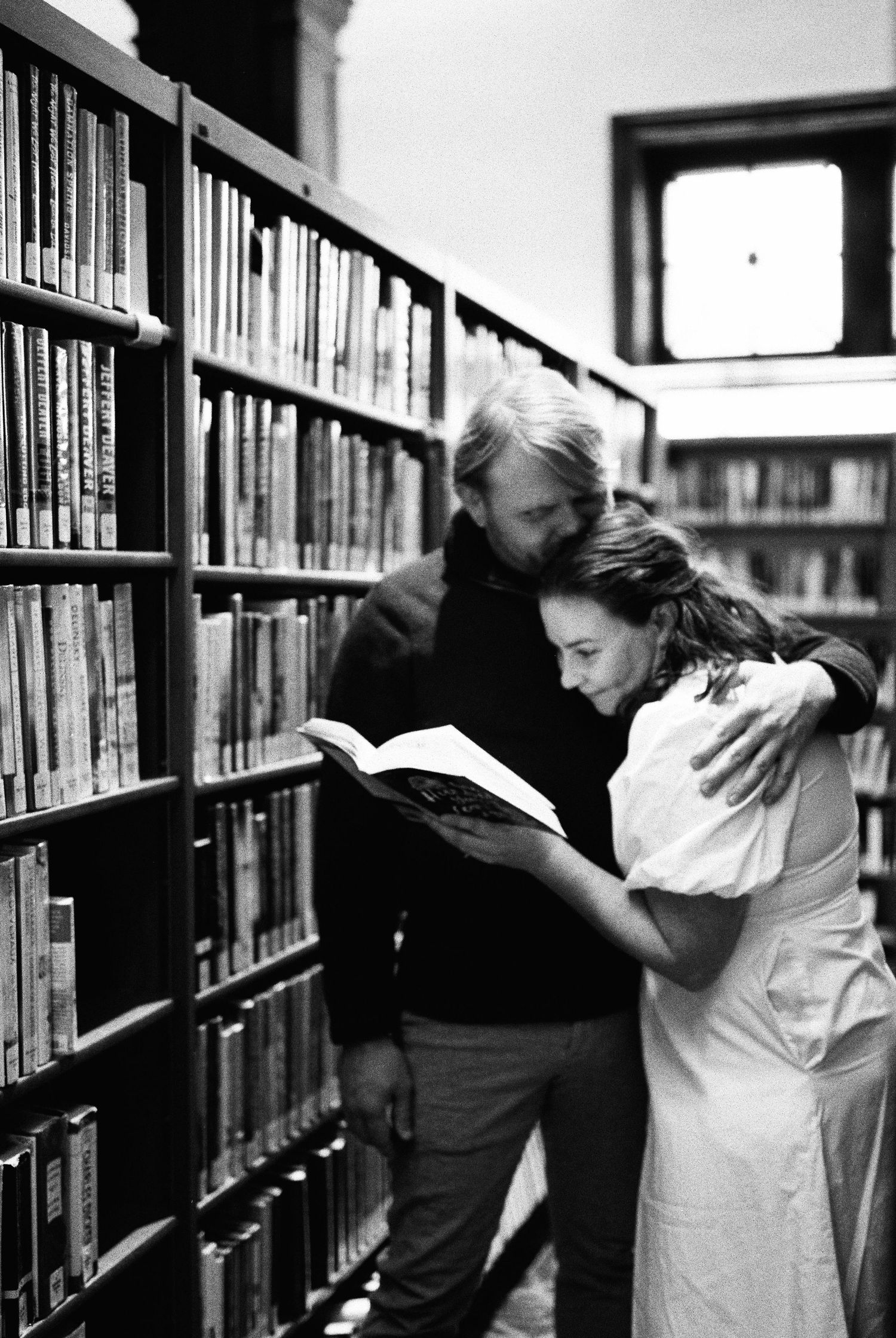 Couple embracing in a library aisle, reading a book together in black and white photo.