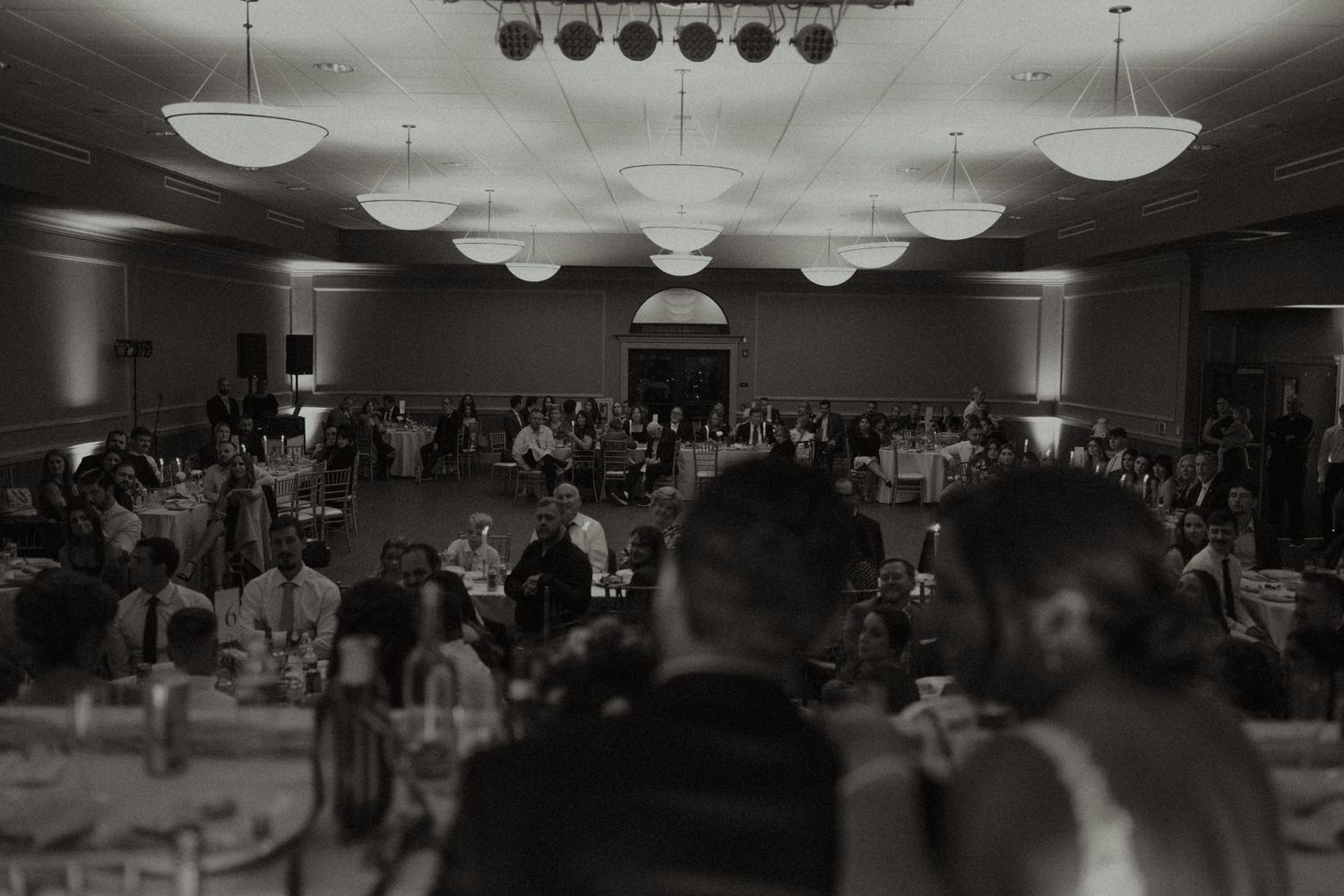 Black and white photo of a large wedding reception hall filled with guests seated at round tables under elegant hanging lights.