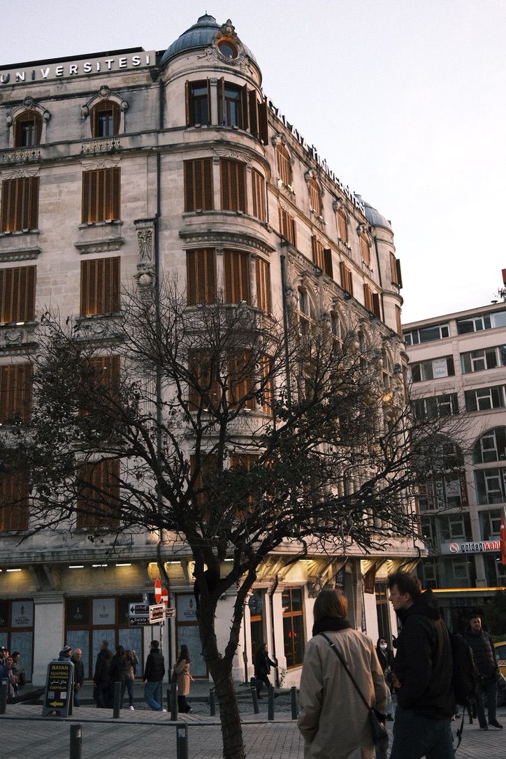 Historic European building with ornate facade, bare winter trees, and pedestrians walking on a cobblestone plaza at dusk.