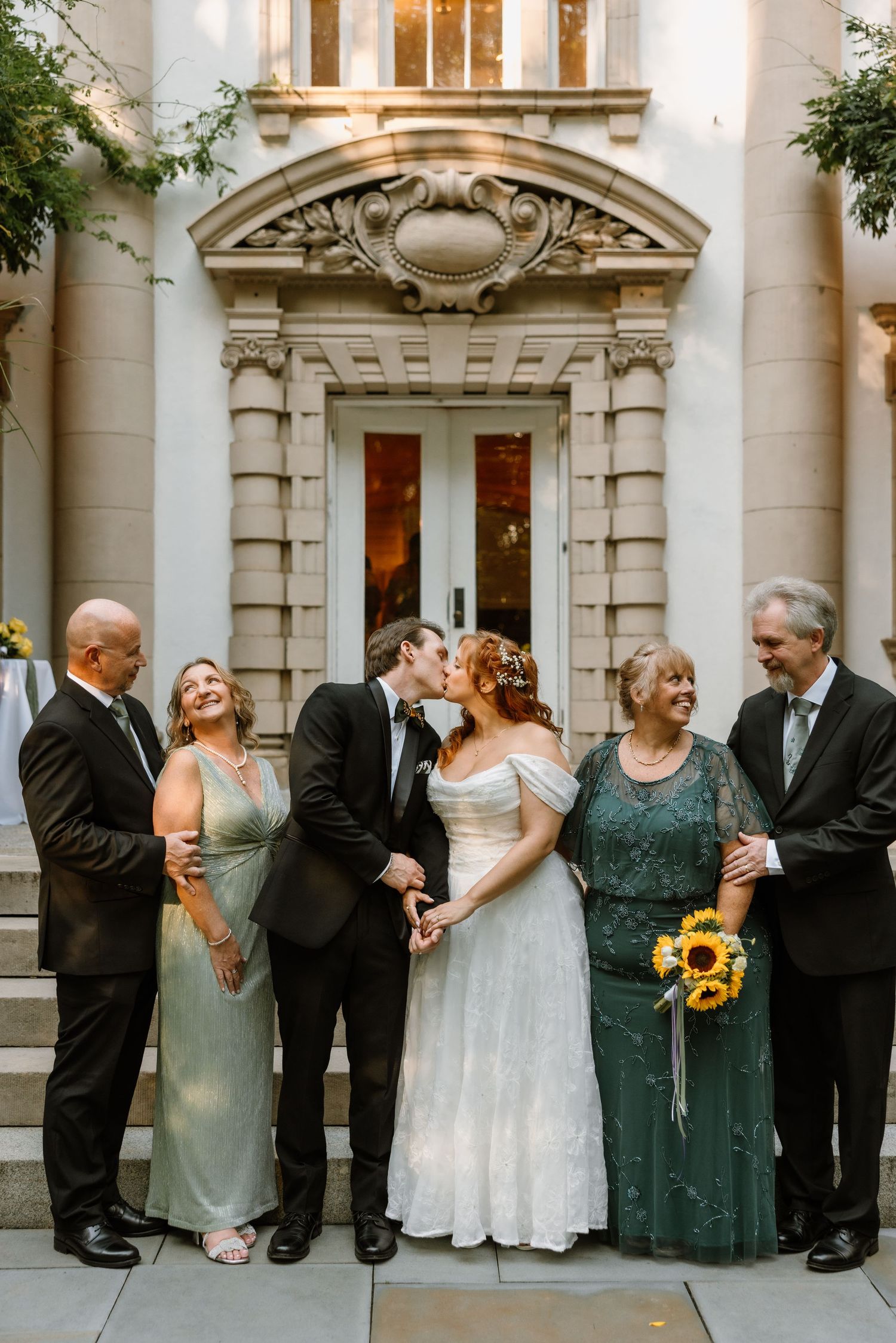 Bride and groom share a kiss flanked by smiling family members outside an elegant stone building with ornate architecture.