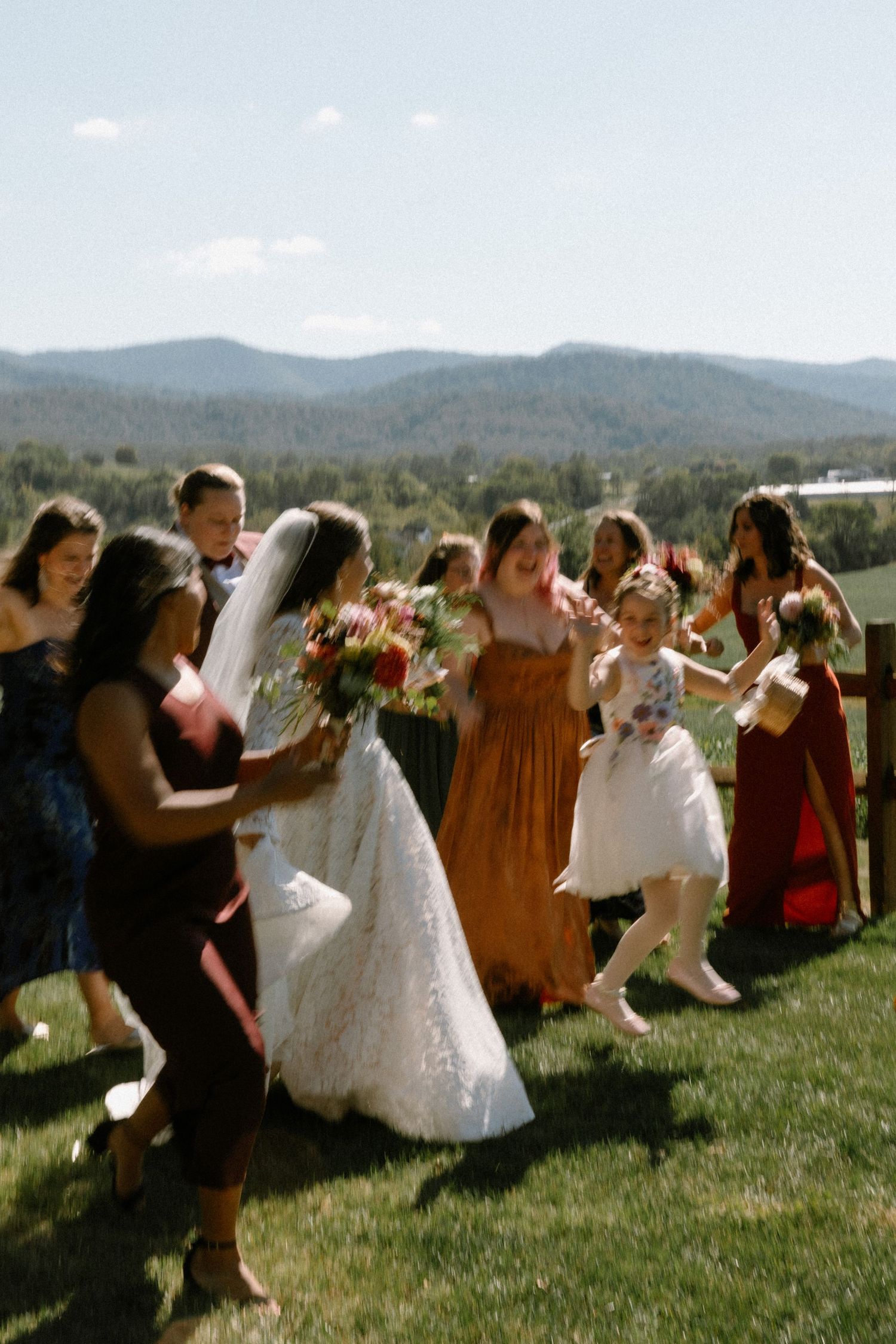Bride in white lace gown walks with bridesmaids in colorful dresses, holding bouquets, with mountain backdrop.