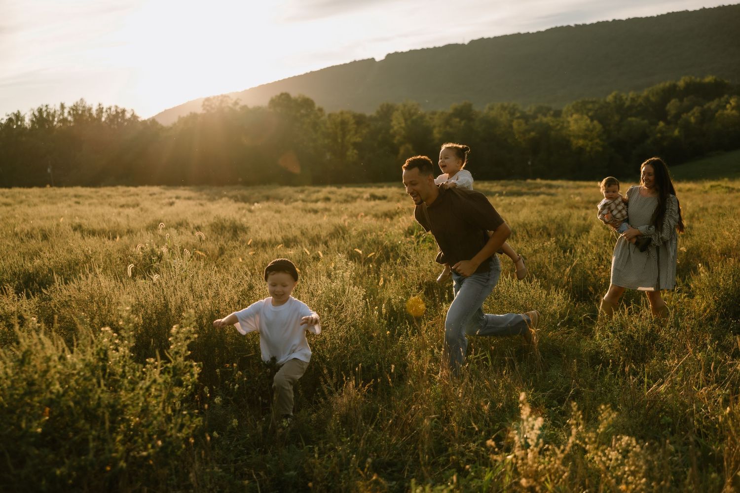 Family playing in a sunlit green field at golden hour with rolling hills and trees in the background.
