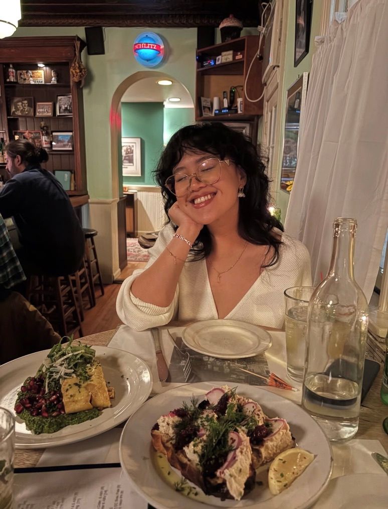 A smiling woman in a white blouse sits at a restaurant table with plates of gourmet food and glasses of wine.