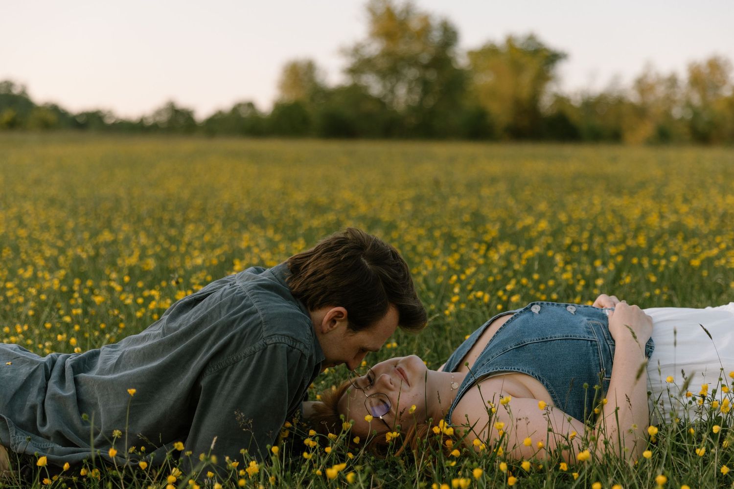 Couple sharing a romantic moment lying in a blooming yellow wildflower field surrounded by lush green trees.