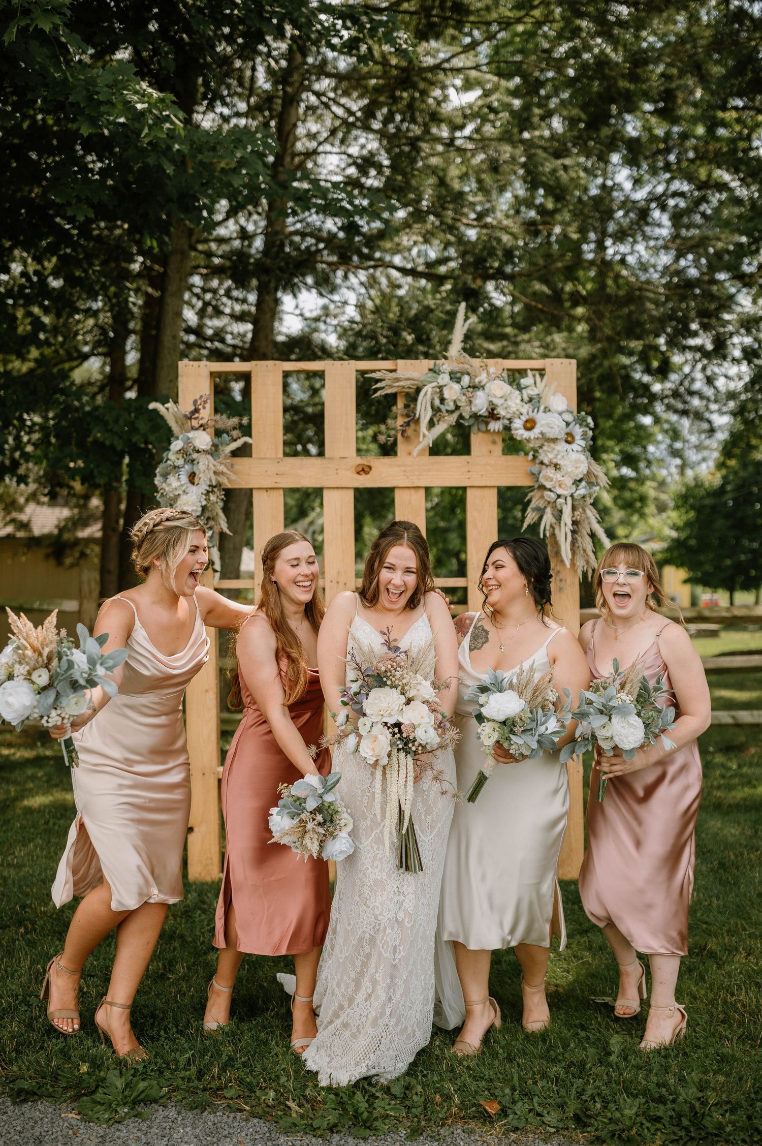 Bride and bridesmaids in mismatched satin dresses laughing outdoors by a wooden arch with floral decor.