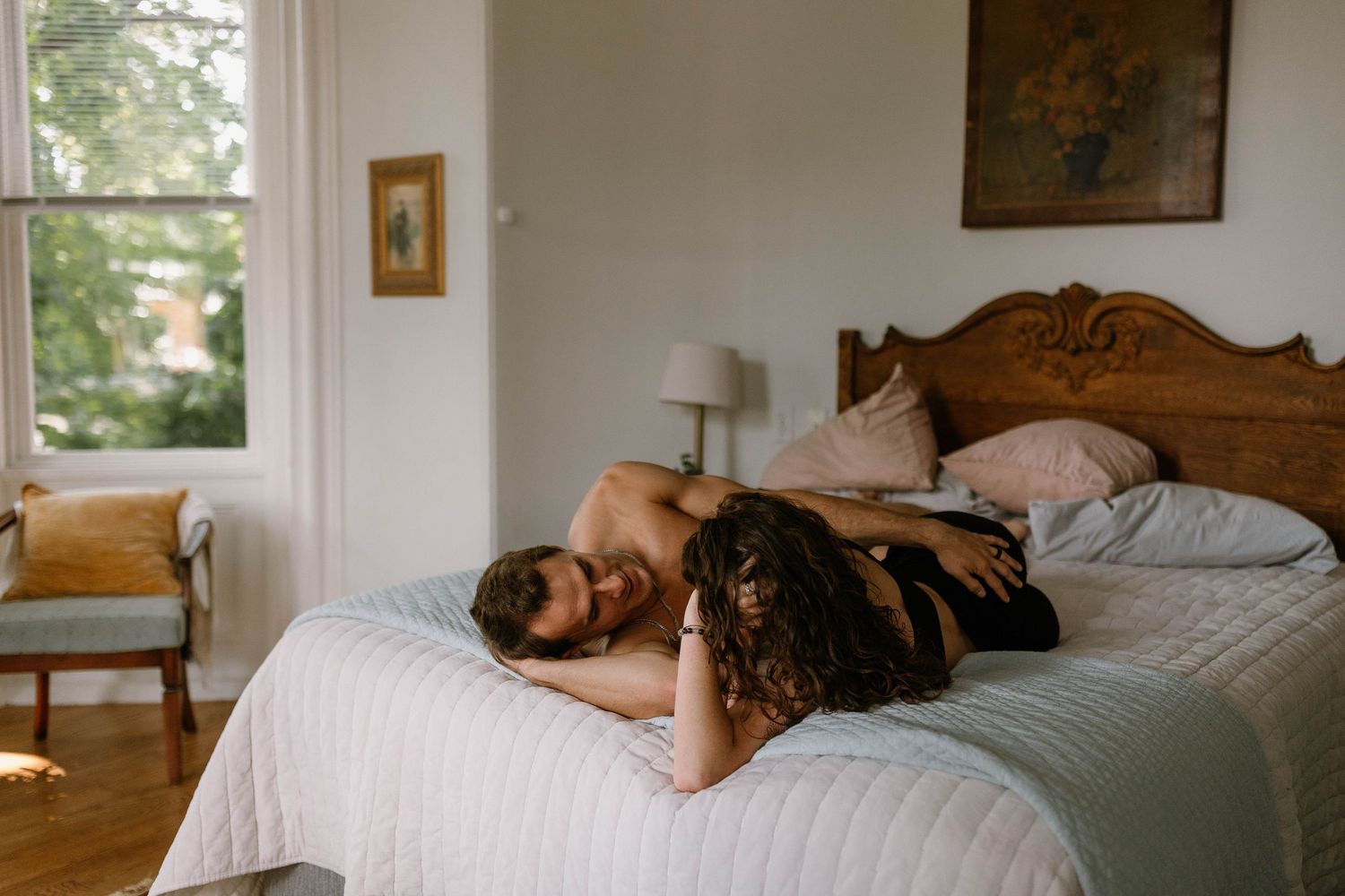 Couple sharing an intimate moment on a bed in a cozy bedroom with natural light and vintage decor.